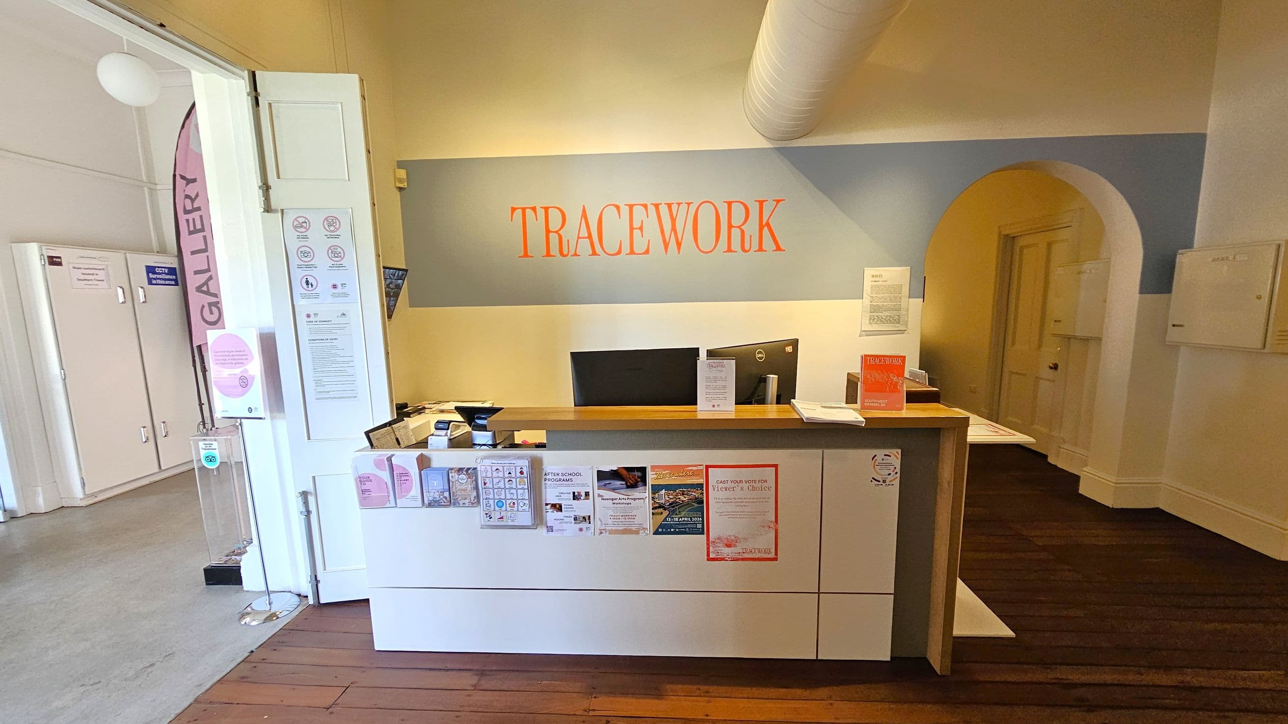 A reception area with a wooden floor. In the centre, a desk with computers, brochures, and notices. The wall behind displays the word "TRACEWORK" in orange. On the left, a doorway with a pink "GALLERY" sign and informational posters. The right side features an arched doorway leading to another room. The ceiling has a visible duct.