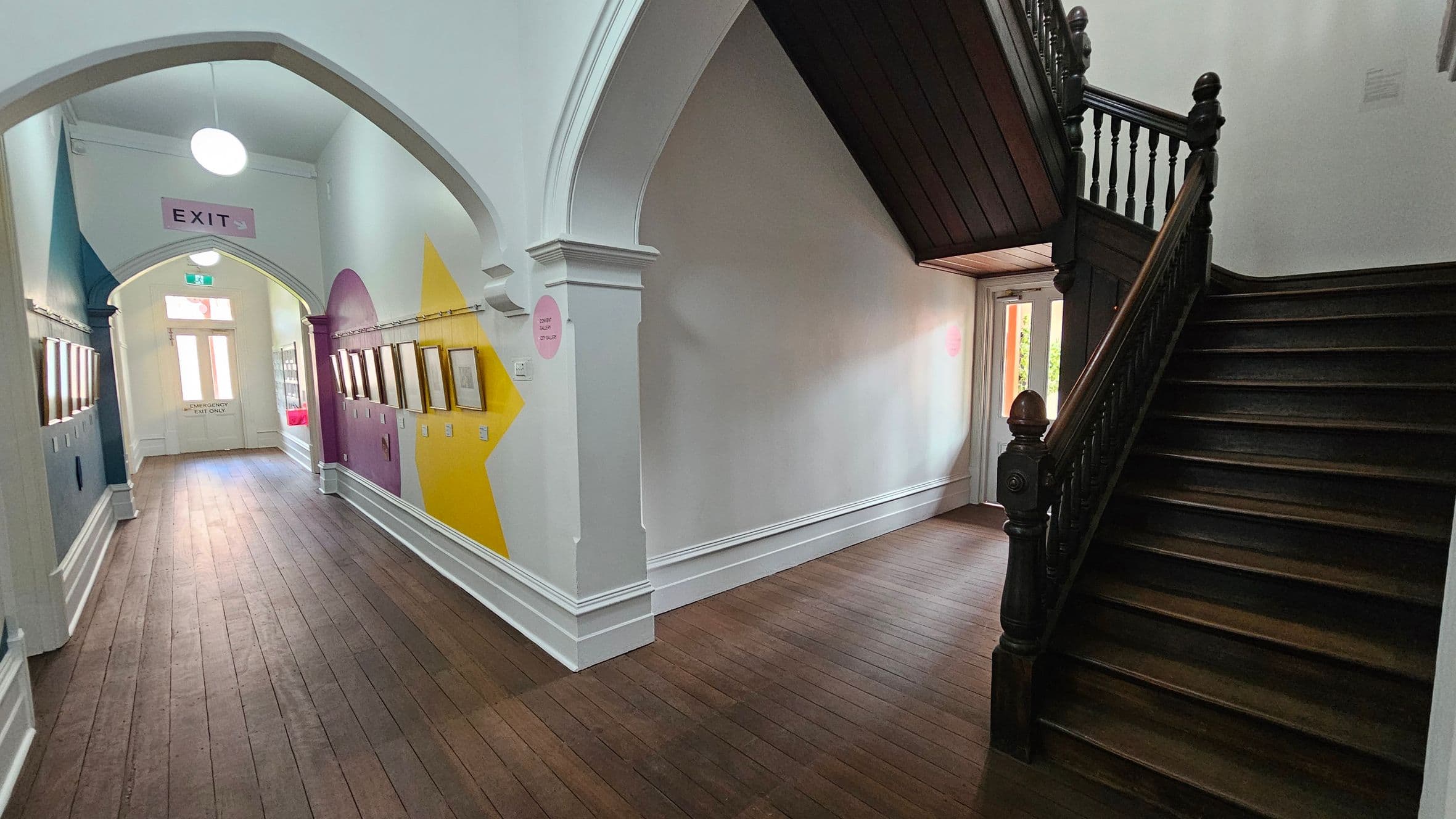 A hallway with wooden flooring and arched ceilings. On the left, colourful geometric patterns adorn the walls, with framed pictures hanging beneath. An "EXIT" sign is visible above a doorway at the end of the corridor. On the right, a dark wooden staircase with ornate railings leads upwards. A small door with glass panels is beneath the staircase. A round ceiling light hangs in the hallway.