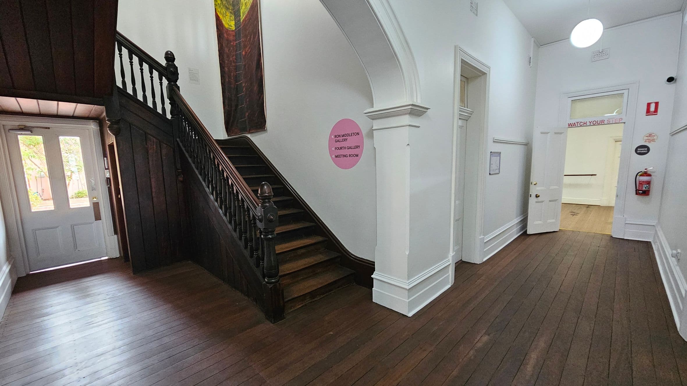 A hallway with wooden flooring and white walls. On the left, a dark wooden staircase with ornate railings leads upward. Below the staircase, a door with glass panels opens to the outside. In the centre, a pink circular sign on the wall indicates directions to various rooms. On the right, an open door with a "WATCH YOUR STEP" sign above it leads to another room. A fire extinguisher is mounted on the wall near the door. The ceiling is white with a round light fixture.