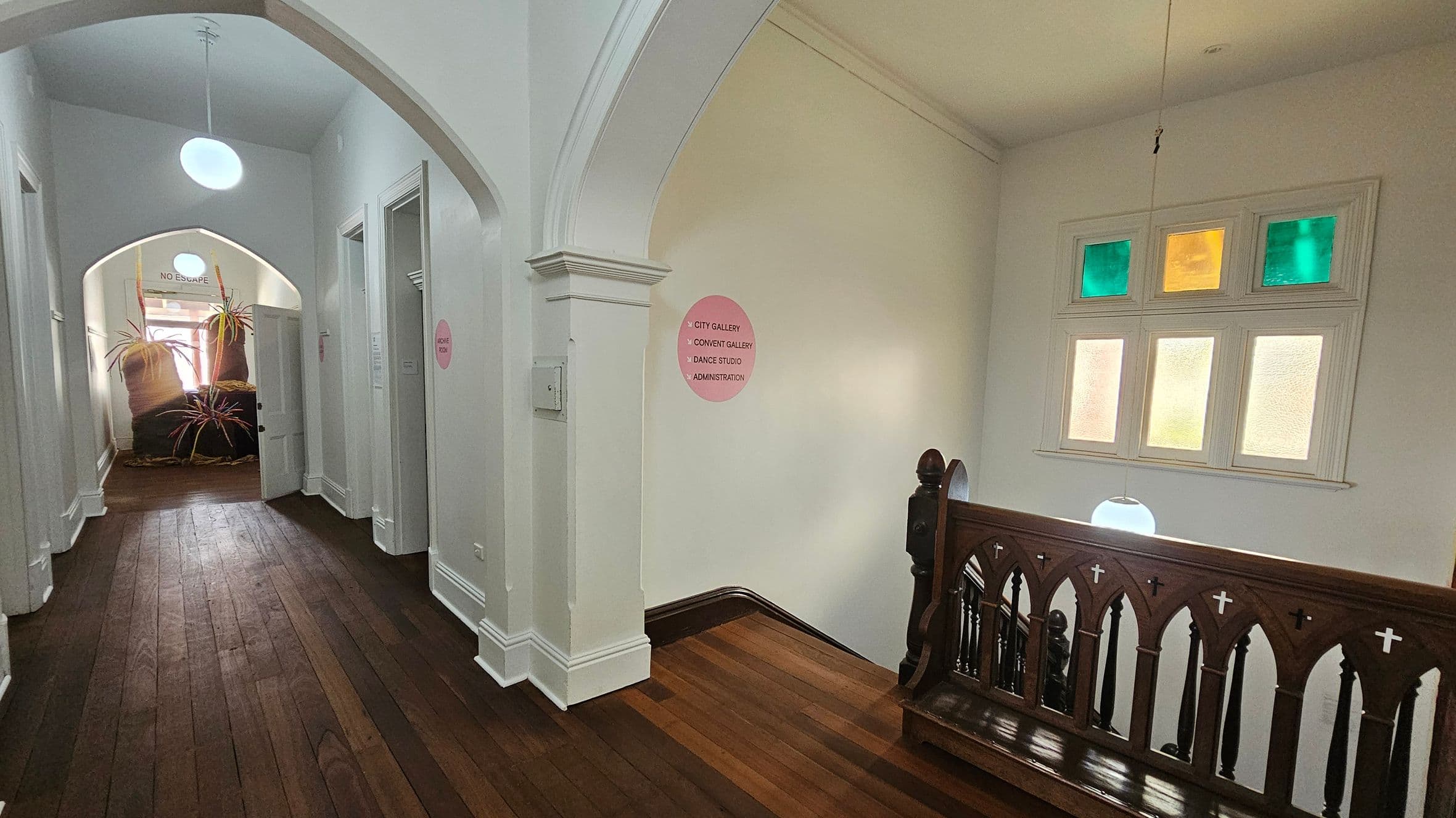 A hallway with wooden flooring and white walls. On the left, there are several doorways and a sign reading "NO ESCAPE" above an open door. At the end of the hallway, large plant sculptures are visible. On the right, a wooden railing with gothic-style arches and crosses overlooks a staircase. Above the railing, a window with coloured panes in green and yellow is present. Two round ceiling lights hang from above, illuminating the space.