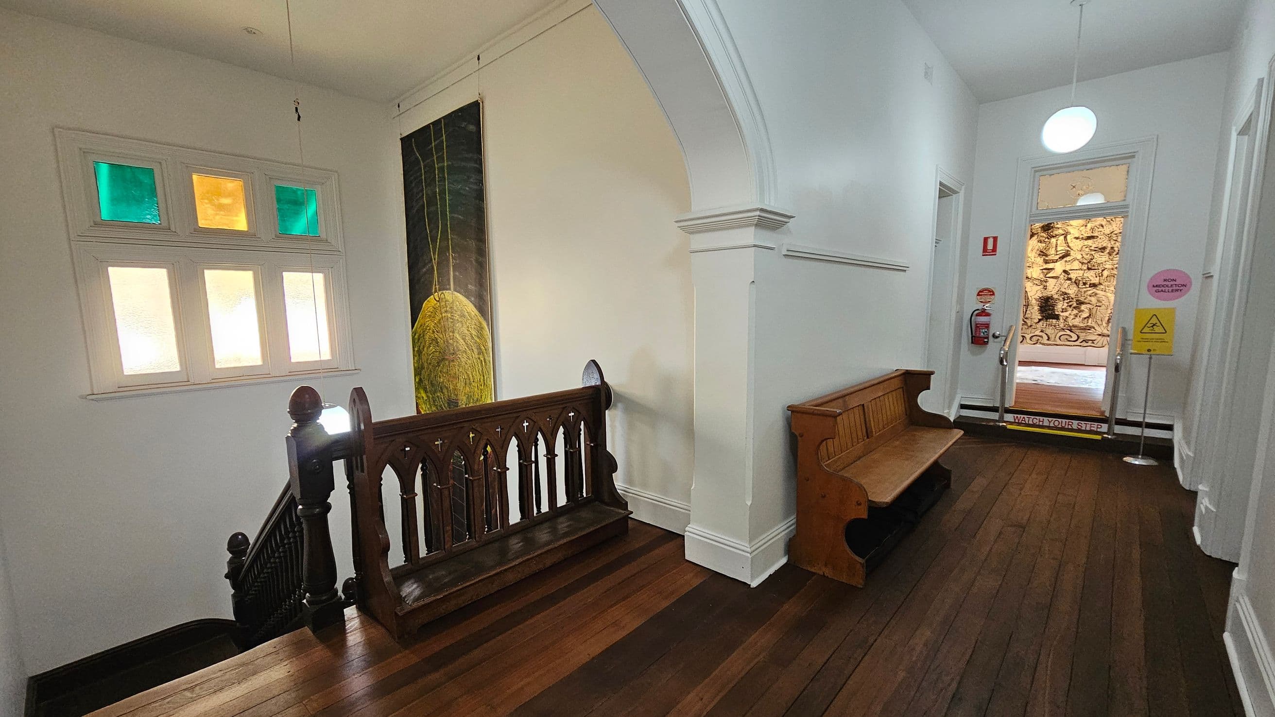 A hallway with wooden flooring and white walls. On the left, a staircase with a dark wooden railing featuring decorative cut-outs. Above the stairs, a window with coloured glass panes in green and yellow. A large painting with a yellow abstract shape hangs on the wall. On the right, a wooden bench is placed against the wall. At the end of the hallway, a door with a glass panel leads to another room, with a "Watch Your Step" sign on the floor and a fire extinguisher nearby.