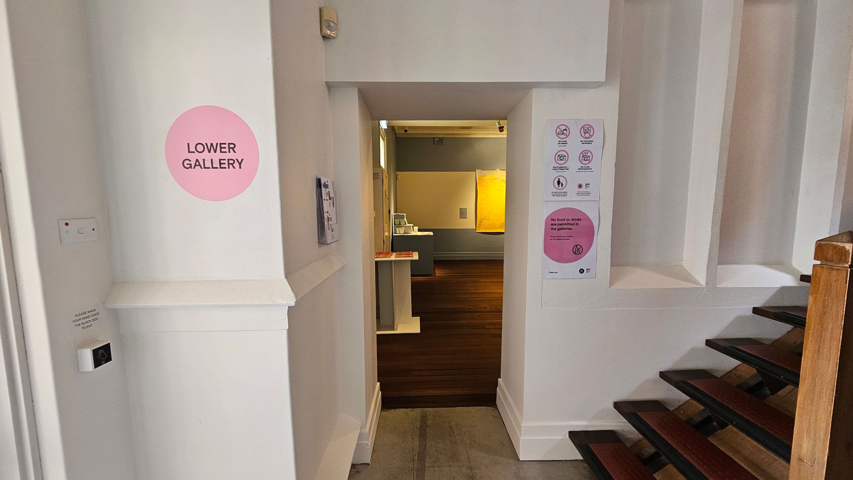 A hallway with a doorway leading to a gallery. On the left, a pink circle sign reads "LOWER GALLERY". A small switch and a notice are below it. On the right, a staircase with wooden steps and a handrail. A sign with gallery rules is posted on the wall. Through the doorway, a room with wooden flooring and a yellow artwork on the wall is visible.