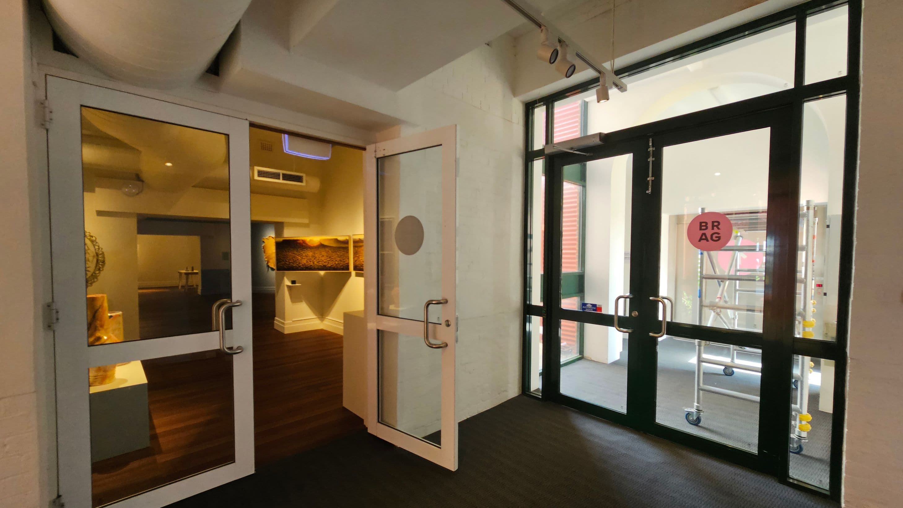 On the left, there is a glass door leading into a room with wooden flooring and artwork on the walls. The room is softly lit, and a sculpture is visible on a pedestal. On the right, there is another glass door with a "BR AG" sign, leading to an area with scaffolding visible outside. The ceiling has exposed pipes and track lighting, and the floor is carpeted near the entrance.