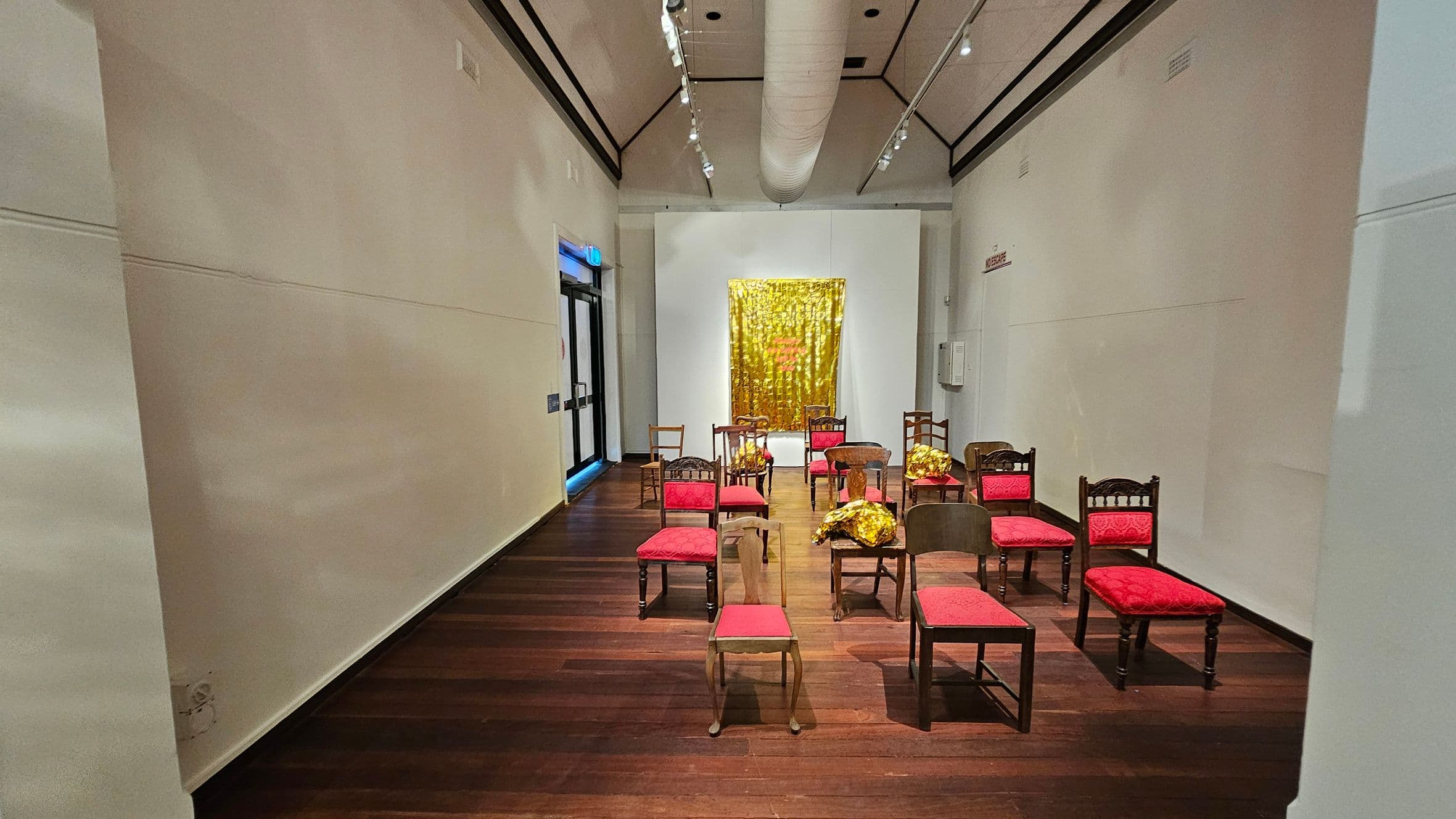 A narrow room with a wooden floor and high ceiling. In the centre, several chairs with red cushions are arranged in rows. Some chairs have gold foil draped over them. At the far end, a large, shiny gold artwork hangs on the wall. The walls are plain and light-coloured. On the left, there is a dark-framed glass door. The ceiling features a large white duct and track lighting.