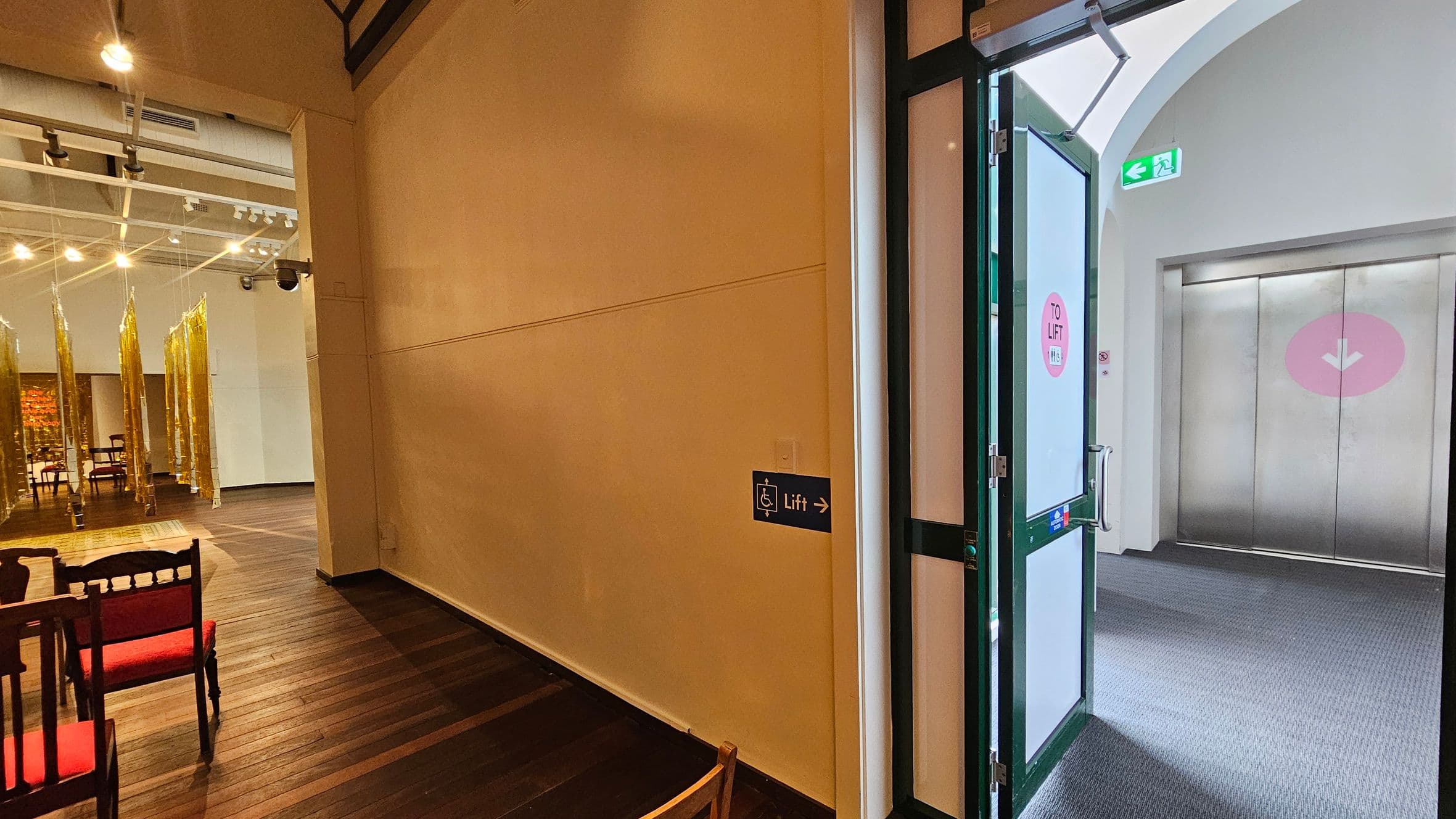 On the right, a door with a sign indicating "TO LIFT" leads to a lift area with a grey carpeted floor and a metal lift door. An exit sign is visible above. On the left, a room with wooden flooring features red-cushioned chairs and gold hanging decorations. The ceiling has exposed beams and spotlights. A sign on the wall points towards the lift.