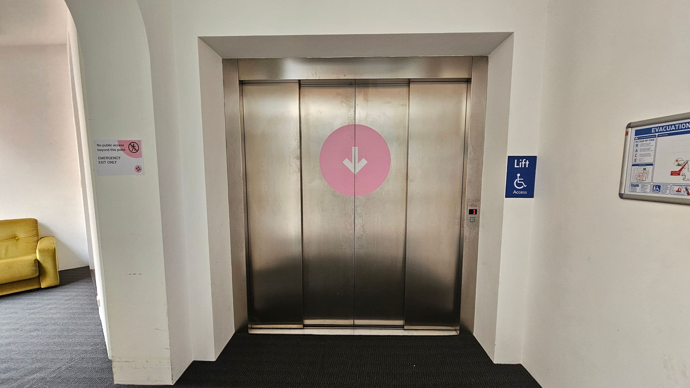 A stainless steel lift with closed doors is centred, featuring a pink circle with a white downward arrow. To the right, a blue sign indicates lift access for disabled individuals, and an evacuation plan is mounted on the wall. On the left, a sign states "No public access beyond this point. Emergency exit only." A yellow armchair is partially visible. The floor is carpeted in dark grey.