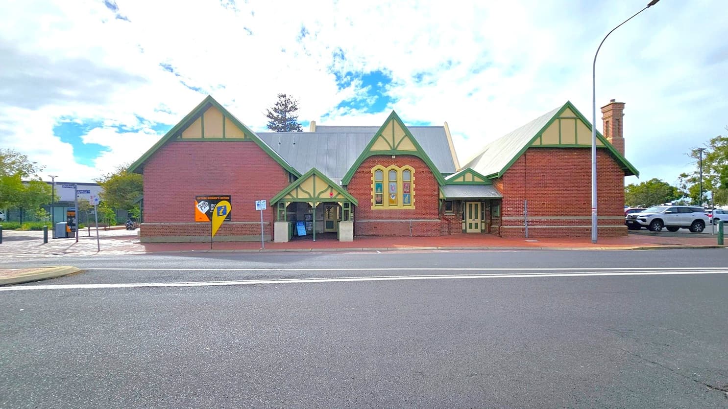 A historic brick building with a gabled roof and green trim stands prominently. It features arched windows and a small entrance with a canopy. A yellow and blue sign is displayed on the left side. The foreground shows a paved road with white lines, and a footpath runs along the building. A streetlamp is positioned to the right, and there are parked cars and trees in the background.