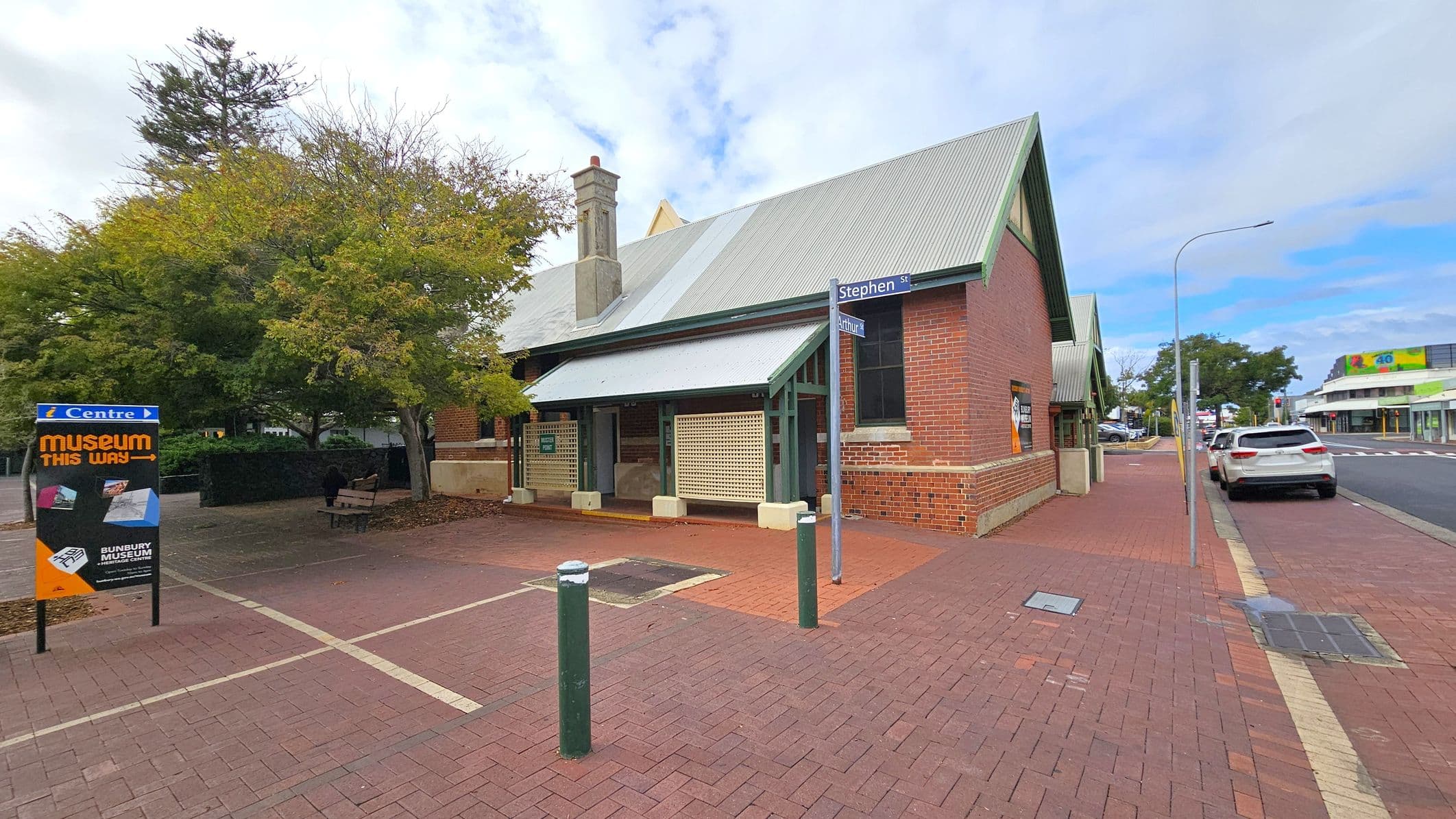 A brick building with a grey metal roof is on the right, featuring a small porch with lattice panels. A street sign indicates "Stephen St" and "Arthur St". On the left, a sign points to a museum, surrounded by trees and a bench. The pavement is red brick, and a white car is parked on the road to the right. The background includes more buildings and a street with a few cars.