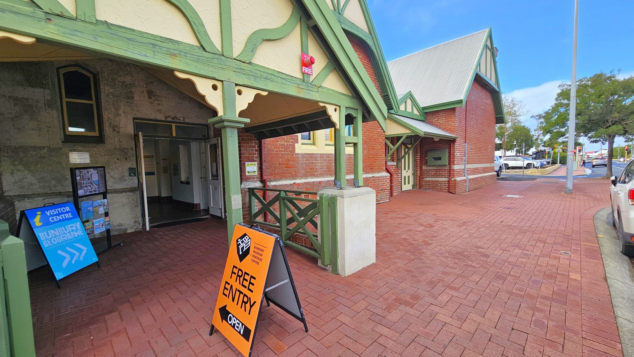 A brick building with green wooden trim and a gabled roof. On the left, there's an entrance with a blue sign for the visitor centre and a map. An orange sign reads "Free Entry" and "Open". The right side shows a continuation of the building with a small porch and a door. The pavement is red brick, and a street with parked cars and a tree is visible in the background.