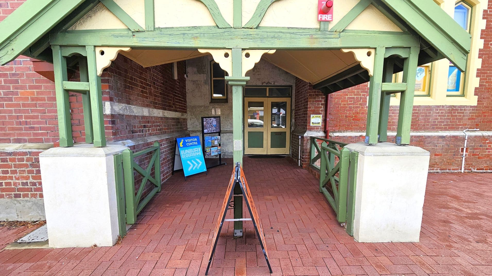 A brick building entrance with a green wooden gabled roof supported by green posts. The floor is paved with red bricks. In the centre, a sandwich board sign stands on the path. To the left, a blue sign indicates a visitor centre, and a rack with brochures is visible. The entrance door is wooden with glass panels. A red fire alarm is mounted above the entrance. The right side shows a window with a yellow frame.