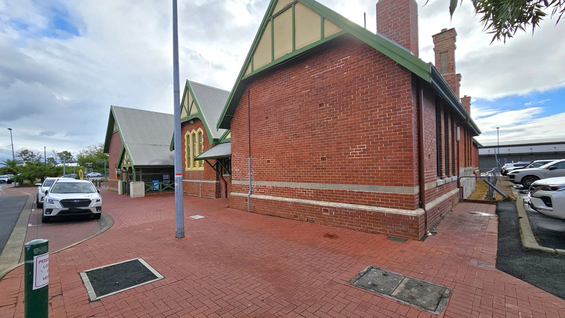 A red brick building with green trim and a gabled roof is on the right. It features tall, narrow windows and a chimney. On the left, a row of parked cars lines the street, including a white vehicle in the foreground. The pavement is made of red bricks, and a tall streetlamp stands near the centre. In the background, there are more parked cars and a low building. The sky is overcast with patches of blue.