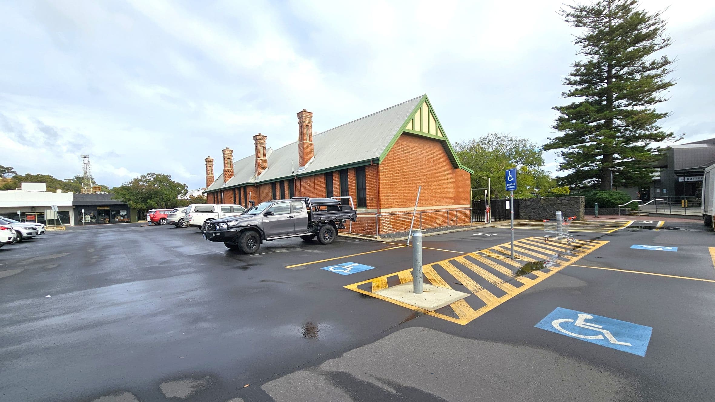 A car park with wet asphalt and marked disabled parking spaces in the foreground. On the right, a brick building with a grey roof and multiple chimneys. A large tree stands nearby. Several vehicles, including a silver pickup truck, are parked. In the background, there are more buildings, including a white one with a shopfront. A shopping trolley is near the parking spaces.