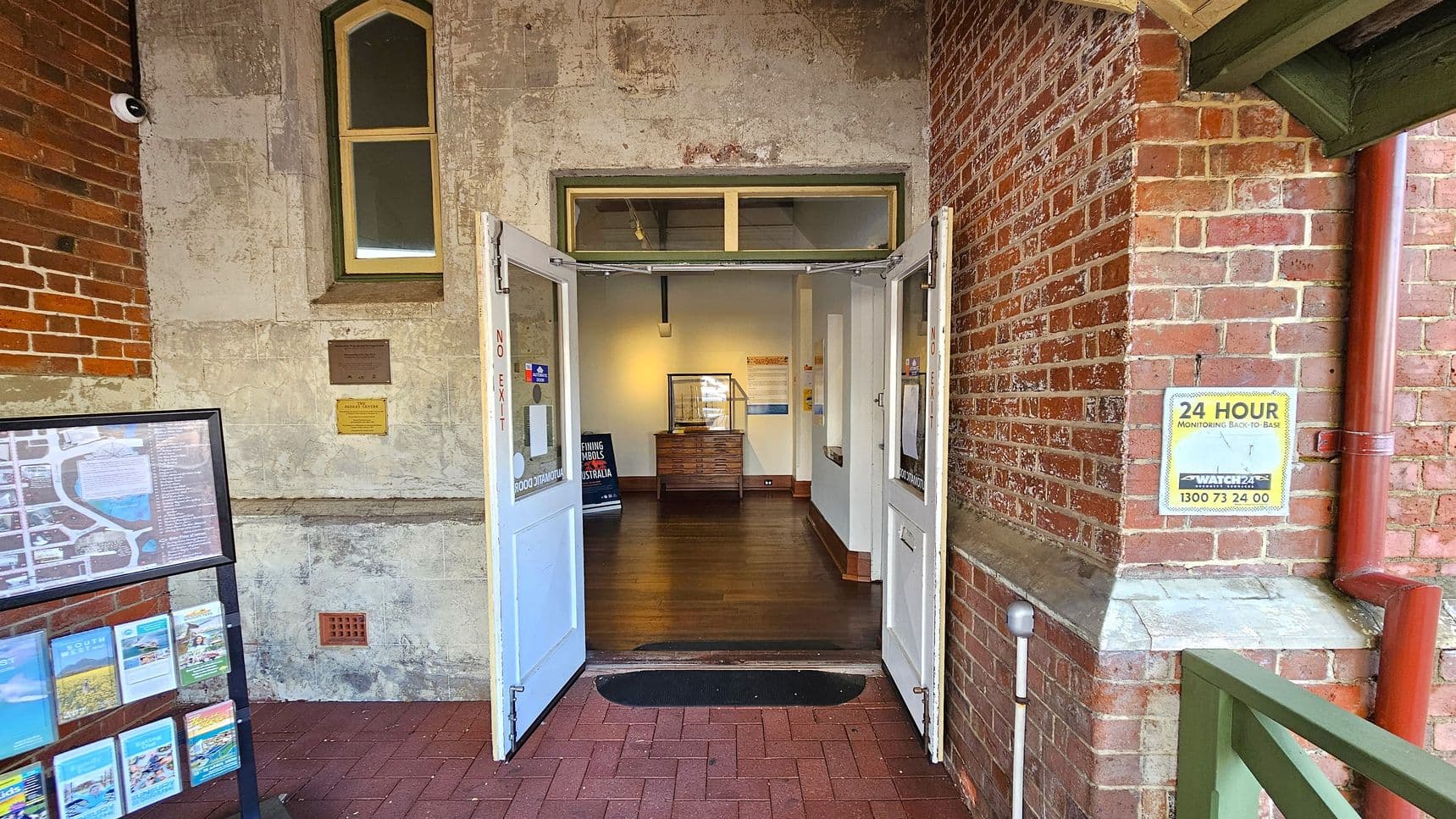 A brick building entrance with double doors open inward. The left wall has a map display stand with brochures and a small window above. The right wall features a security sign and a red drainpipe. Inside, a wooden floor leads to a display case and a sign. The walls are a mix of exposed brick and plaster. A security camera is mounted on the left wall.