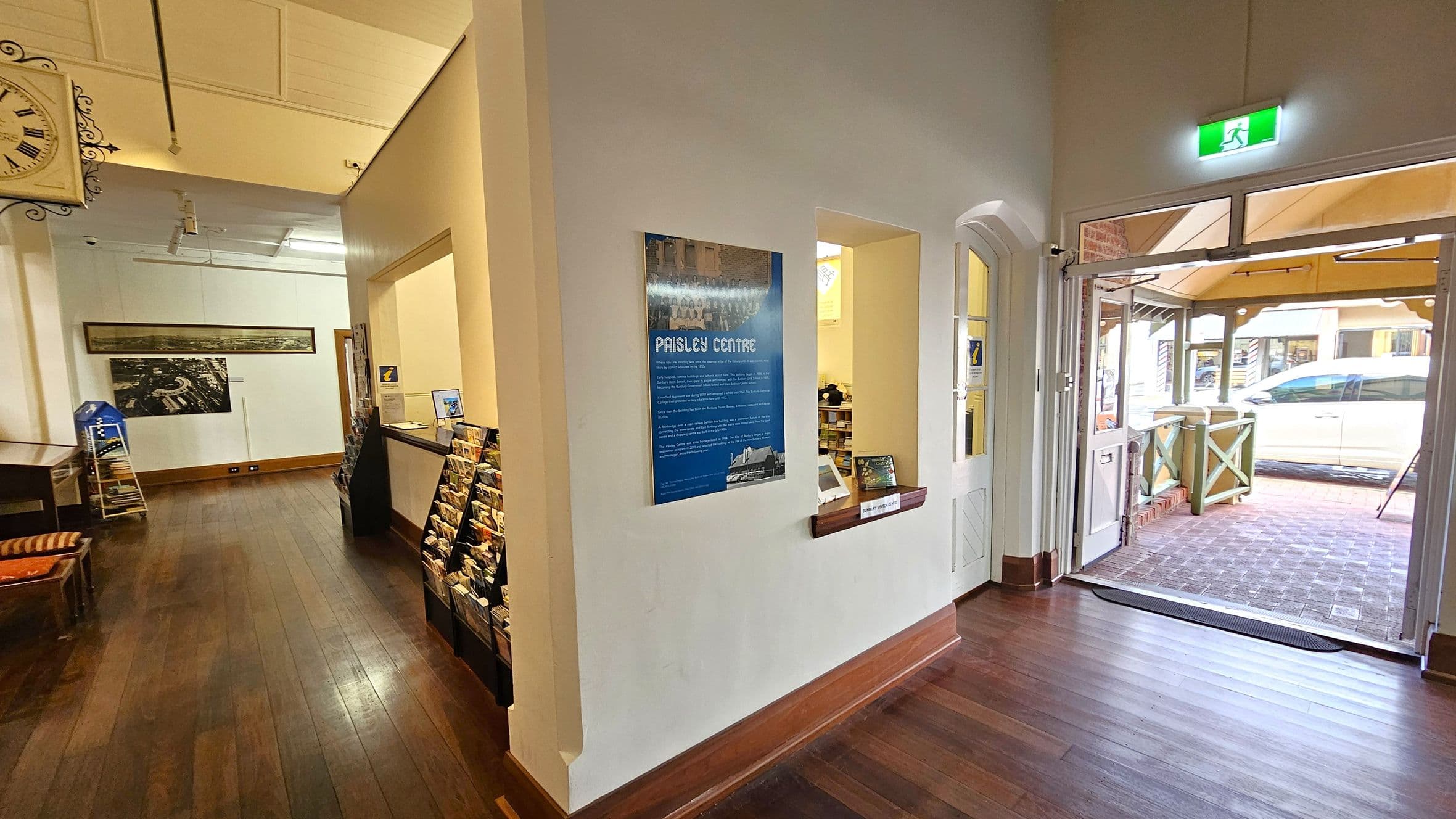 A room with wooden flooring and a high ceiling. On the left, there's a wall-mounted clock and a display rack with brochures. A large photograph is mounted on the wall. In the centre, a blue sign reads "Paisley Centre" next to a windowed counter. On the right, a glass door leads outside, where a white car is visible. An illuminated exit sign is above the door.