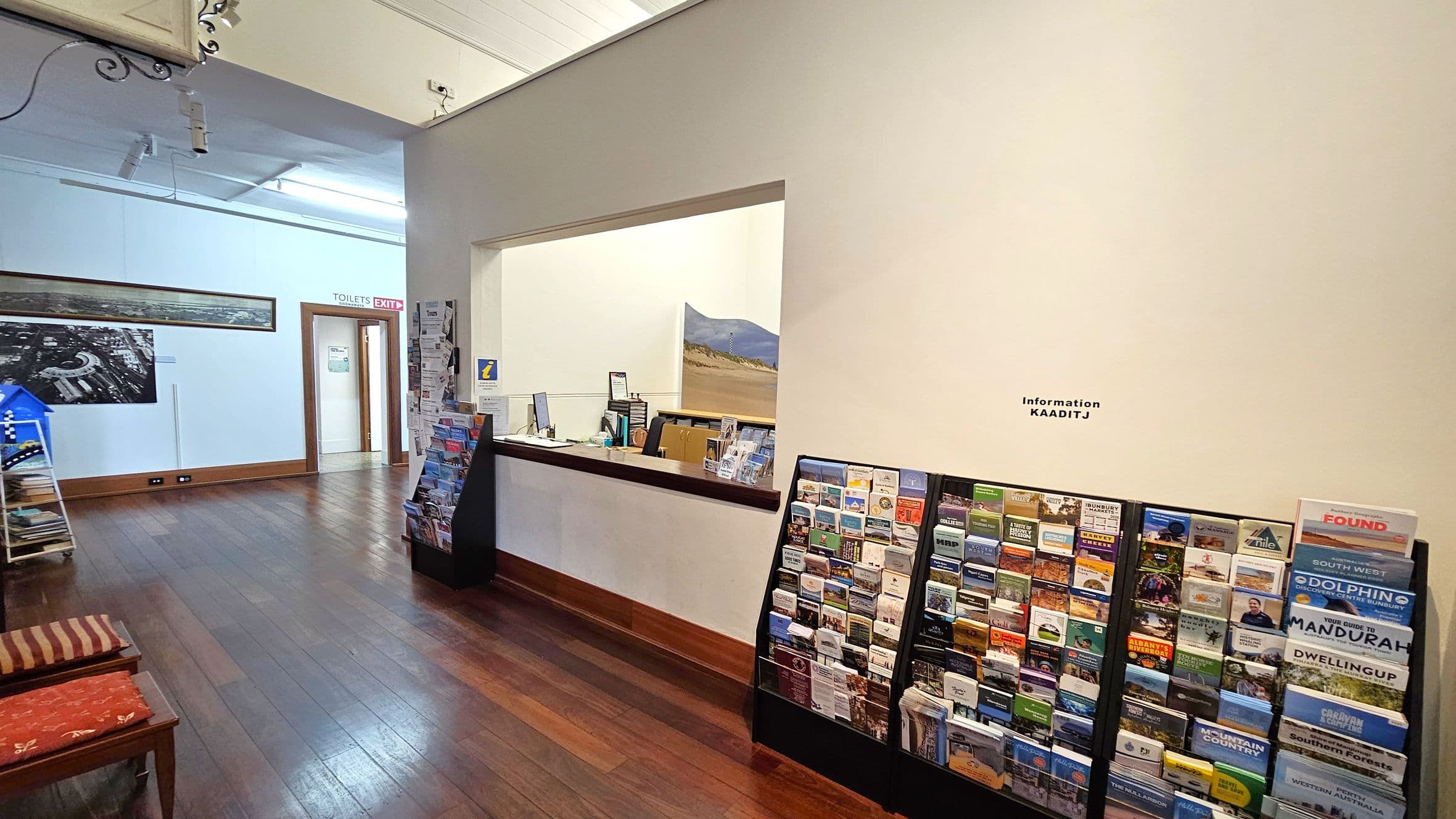A room with wooden flooring and a white ceiling. On the right, there is a wall with a large window opening into an information desk area, featuring brochures and pamphlets displayed on racks. The left side has a doorway marked "TOILETS EXIT" and a large black-and-white photograph on the wall. Two chairs with striped cushions are visible in the foreground. The lighting is provided by ceiling-mounted fixtures.