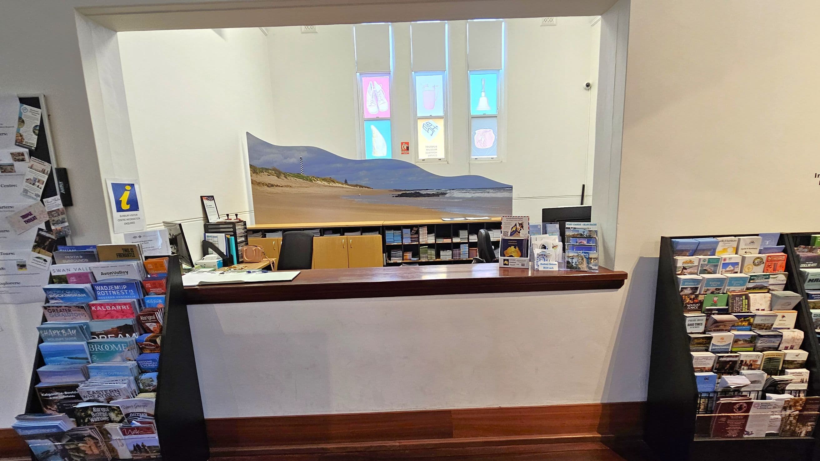 A reception area with a wooden counter in the foreground. On the left and right, there are racks filled with brochures and pamphlets. Behind the counter, a large mural of a beach scene is visible. Above the mural, three vertical windows display colourful designs. The floor is wooden, and the walls are light-coloured. Various informational materials and a small sign with an "i" for information are on the counter.