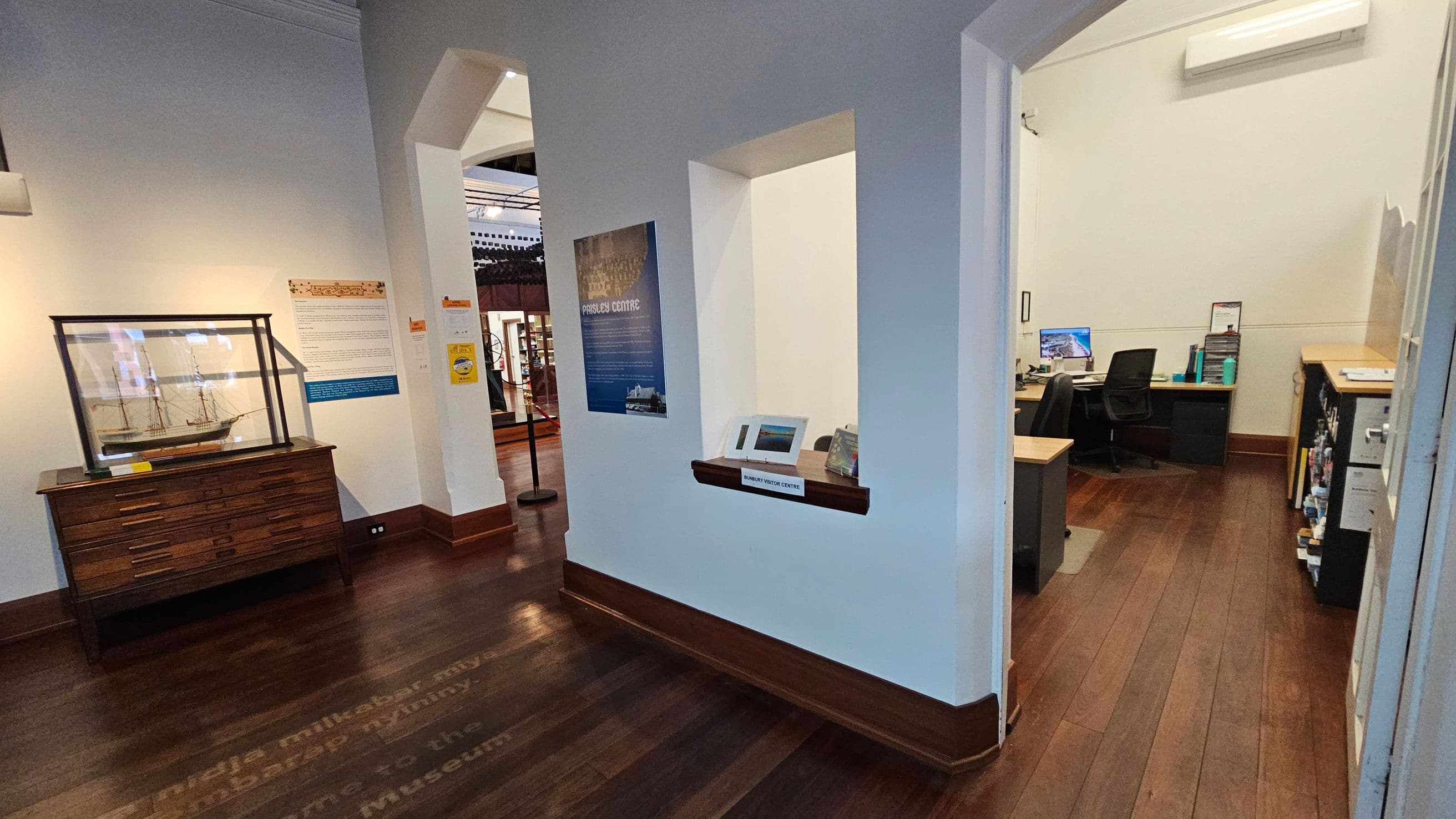 A room with wooden flooring and white walls. On the left, a model ship is displayed in a glass case atop a wooden cabinet. Adjacent to it, informational posters are mounted on the wall. In the centre, a small window labelled "Railway Visitor Centre" is set into the wall. On the right, an office space is visible through an open doorway, featuring a desk with a computer, chairs, and shelves with books and files. An air conditioning unit is mounted on the wall above.