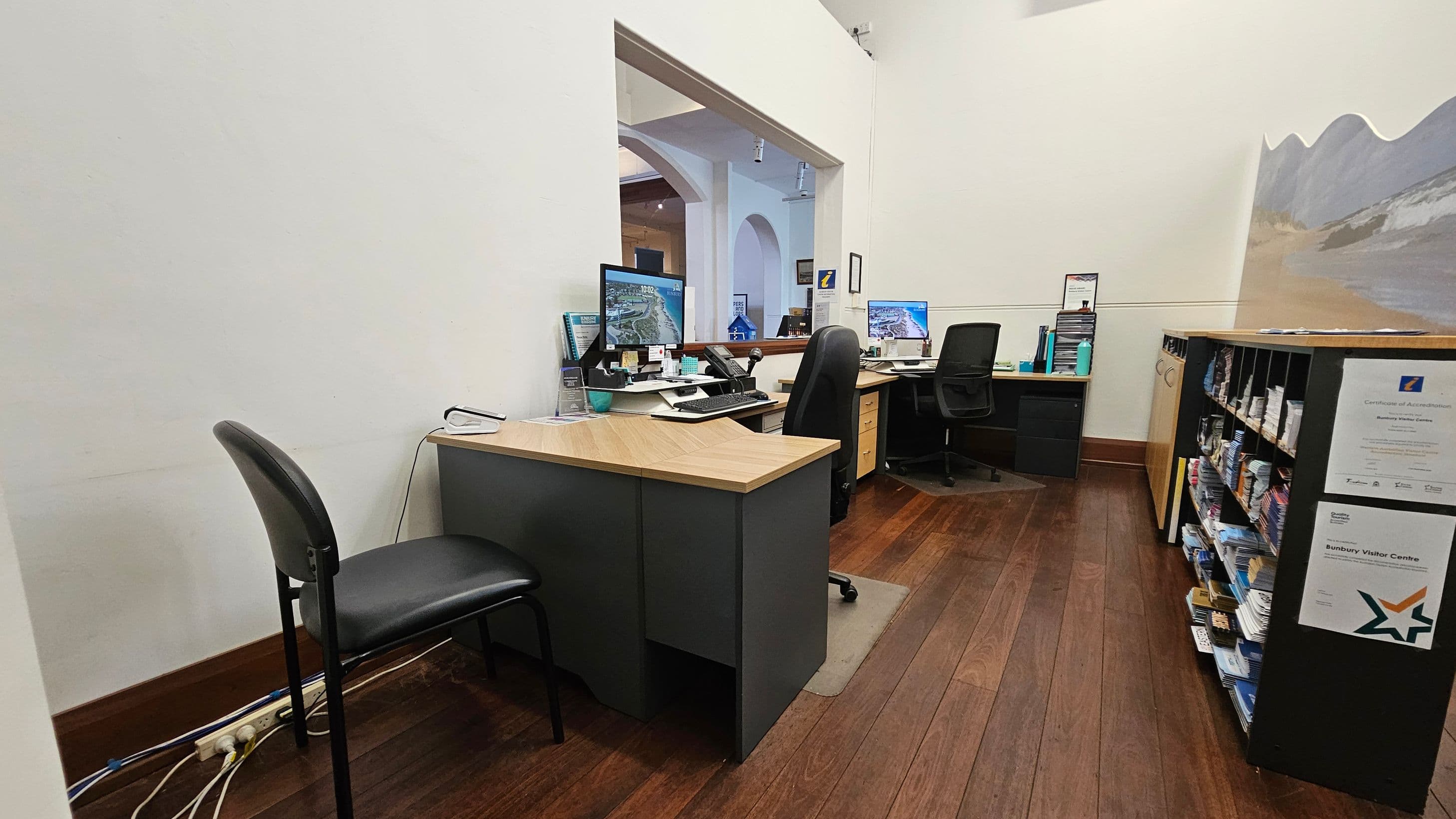 An office space with wooden flooring. On the left, a black chair is positioned near a desk with a computer monitor, keyboard, and phone. Cables are visible on the floor. In the centre, another desk with a computer and office chair is visible through an open partition. On the right, a shelving unit holds brochures and papers, with certificates displayed on the side. The walls are light-coloured, and a mural or painting is partially visible on the right wall.