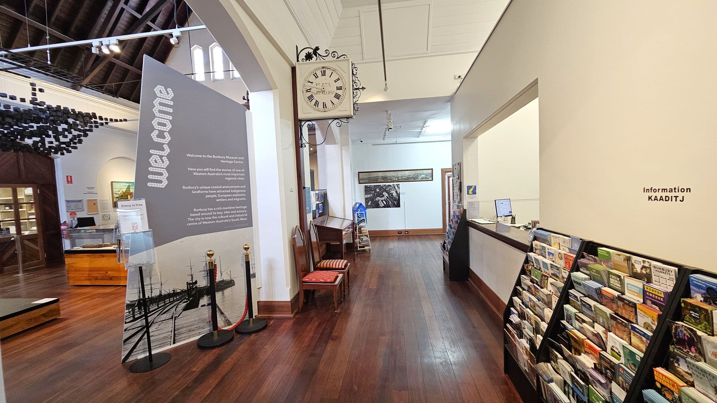 A museum interior with a wooden floor and high, dark wooden ceiling beams. On the left, a large welcome sign with text and images stands near a wooden counter. Behind it, a display area with various exhibits. In the centre, a vintage clock is mounted on the wall above two chairs. On the right, a wall with a window opening to an information desk, and a rack filled with brochures. The space is well-lit with track lighting.