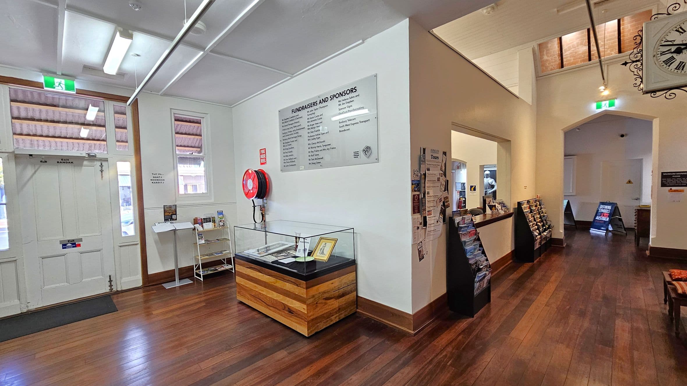 A room with wooden flooring and a white ceiling with fluorescent lights. On the left, there is a white double door with an exit sign above it, and two windows. A display case with a wooden base is against the wall, along with a fire hose reel and a sign listing fundraisers and sponsors. In the centre, there is a small shelf with brochures. On the right, a hallway leads to another area, with racks of brochures and a large wall clock. An open doorway reveals more signage and a lit area beyond.
