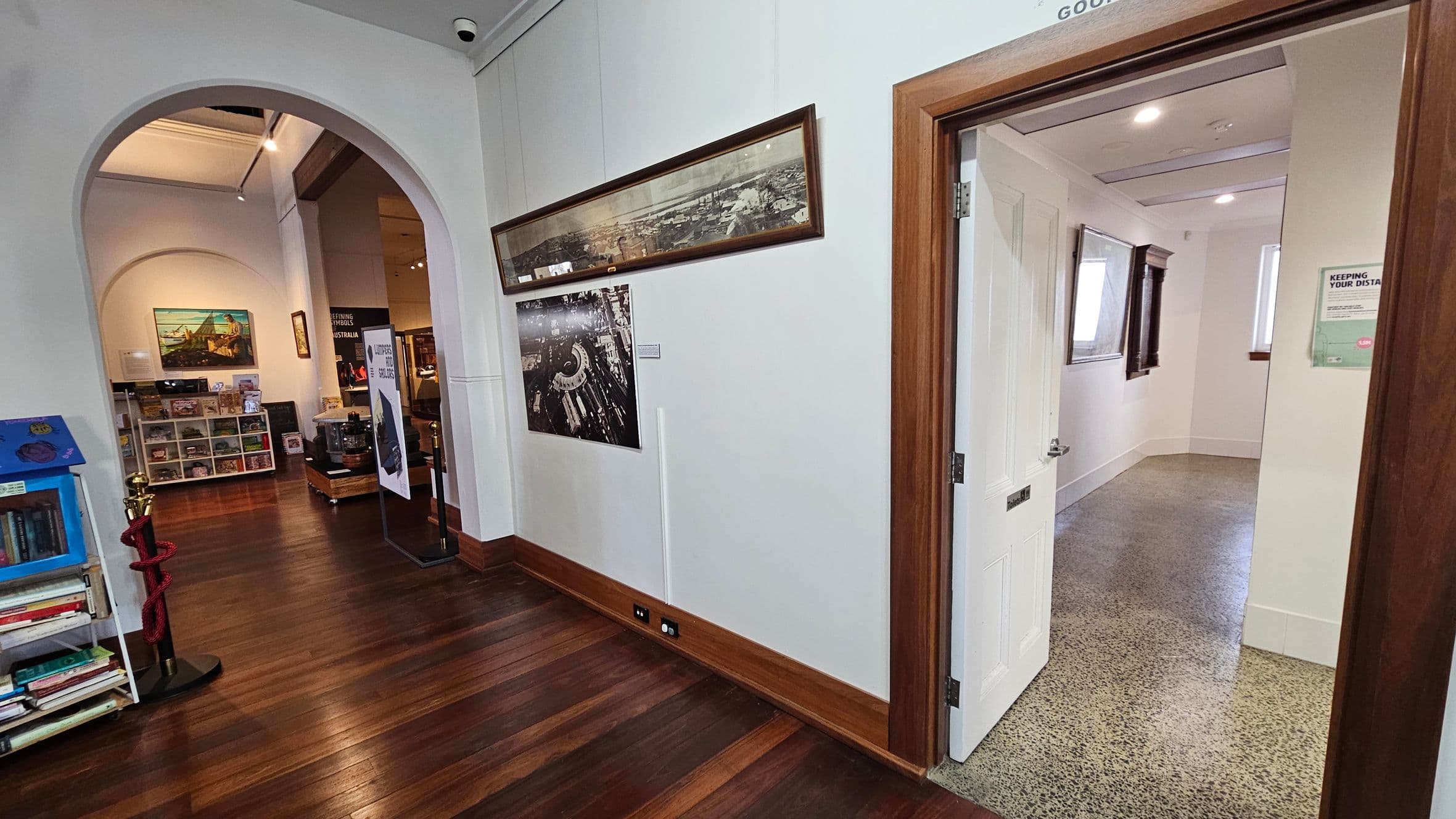 A room with wooden flooring and white walls. On the left, there's an arched doorway leading to another room with bookshelves and a painting on the wall. A red rope barrier is visible near a blue shelf filled with books. On the right, an open door leads to a smaller room with a speckled floor and windows. Two framed pictures hang on the wall near the door. The ceiling has recessed lighting.