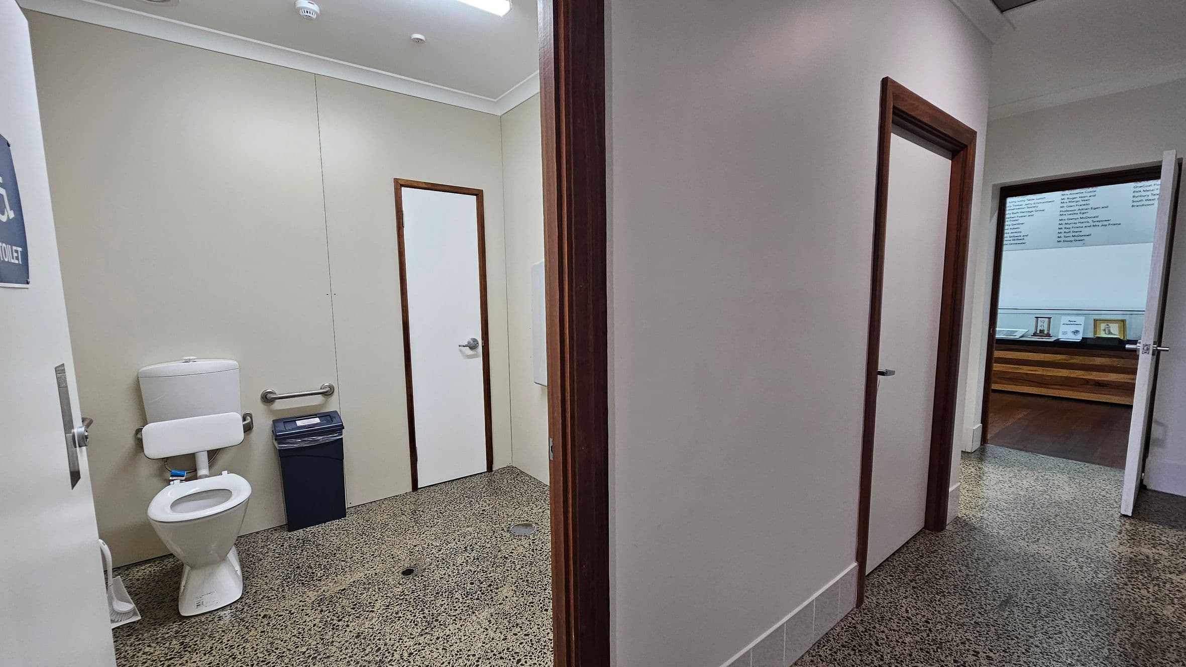 On the left, a bathroom with a white toilet, a grab bar, and a bin against a light-coloured wall. The floor is speckled with green and brown tones. In the middle, a doorway with a wooden frame leads to another room. On the right, another open door reveals a room with a wooden counter and framed items on display. The ceiling is white with a light fixture.