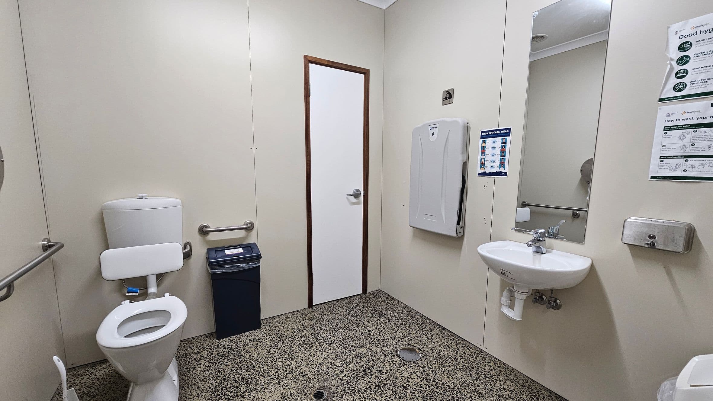 A public restroom with a speckled floor. On the left, there is a toilet with a support bar and a bin nearby. In the centre, a white door with a metal handle. On the right, a baby changing station is mounted on the wall, with a mirror above a small sink. A soap dispenser is fixed to the wall next to hygiene instruction posters. The walls are plain, and the ceiling is white with a light fixture.