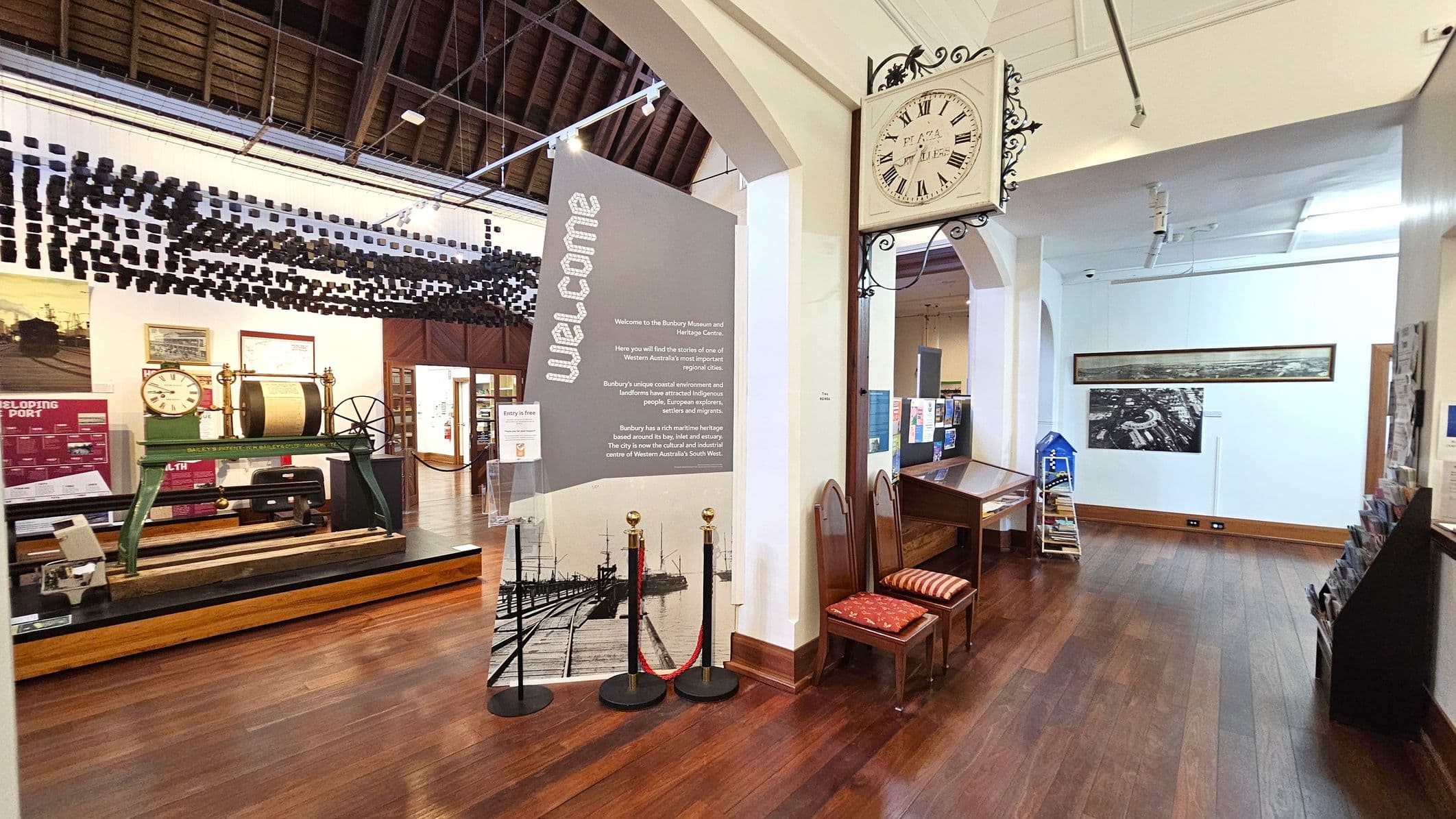 A museum interior with wooden flooring and a high, exposed wooden ceiling. On the left, there is a display featuring a vintage clock and machinery. A large welcome sign stands nearby. On the right, a wall-mounted clock with Roman numerals is above a wooden bench with red cushions. Brochures are displayed in a rack, and a panoramic photograph is mounted on the wall. The space is well-lit with track lighting.