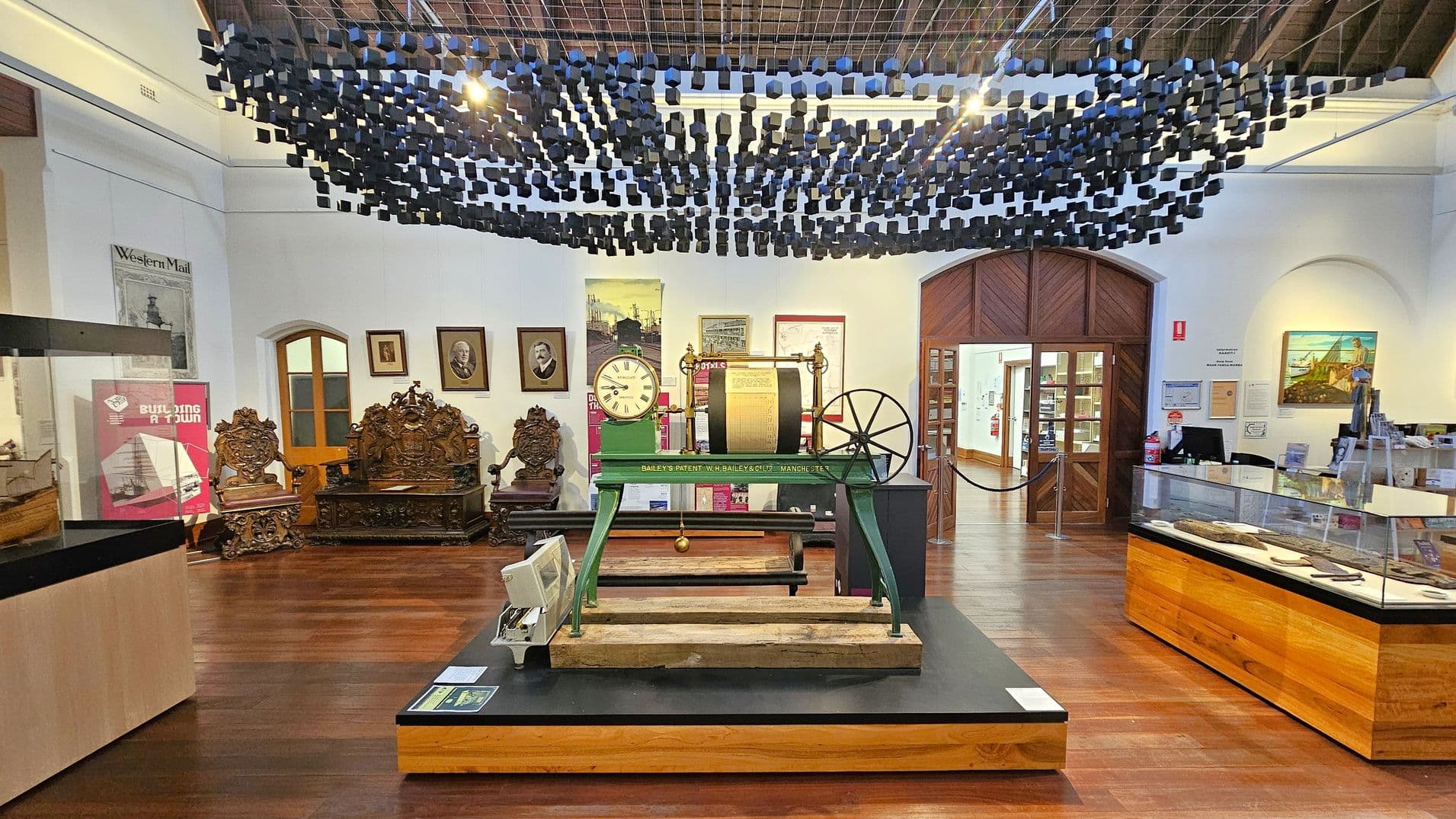 A museum interior with a wooden floor and a ceiling adorned with hanging black cubes. In the centre, there's a large green machine with a clock and a wheel, labelled "Bailey's Patent." Behind it, ornate wooden chairs and a table are displayed. The walls feature framed portraits and posters. On the right, a glass display case contains various artefacts. A wooden door with glass panels is in the background, leading to another room.