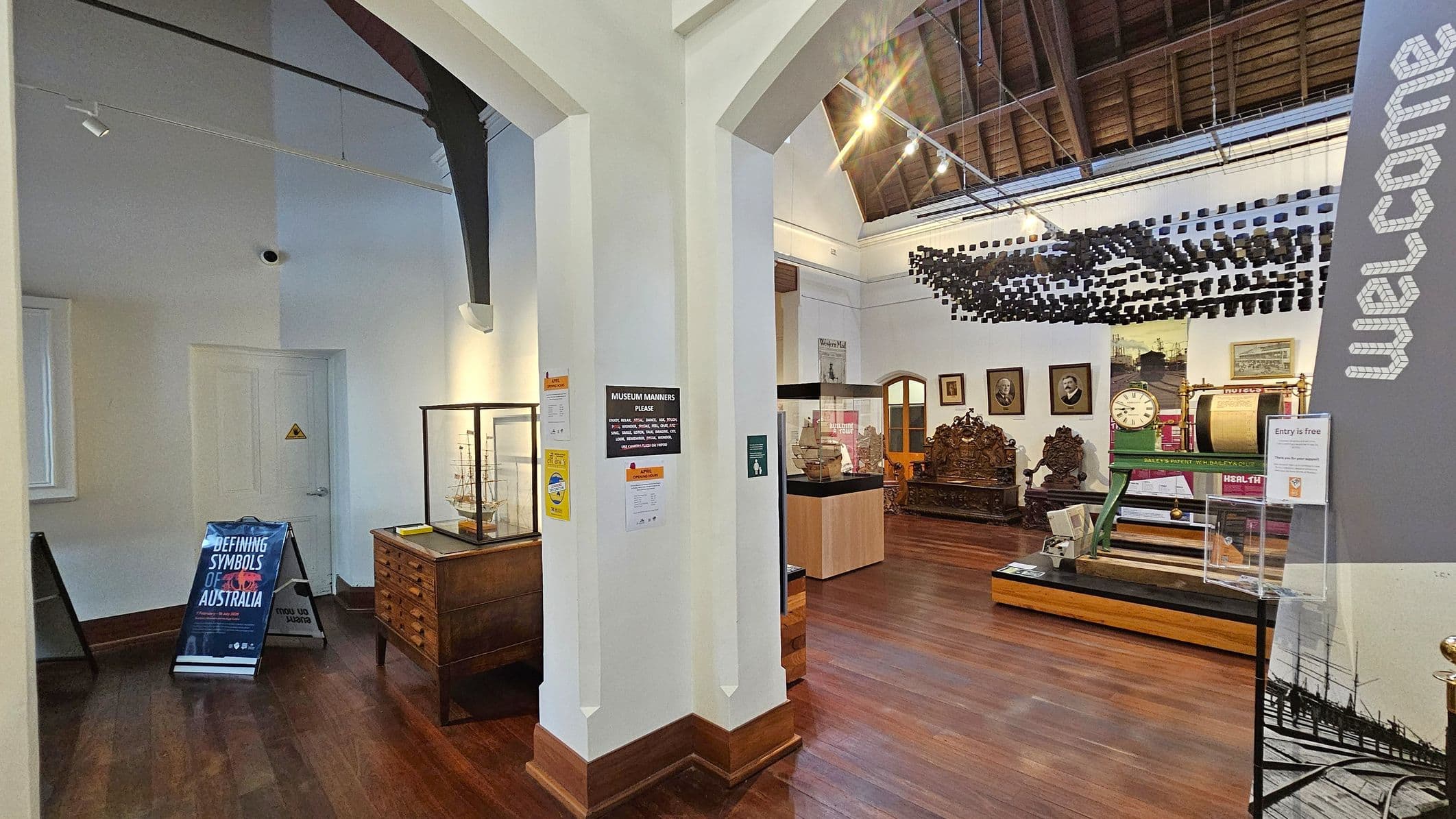 Museum interior with wooden floors and a high, wooden-beamed ceiling. In the centre, a white pillar displays signs about museum etiquette. To the left, a glass display case with a model ship sits on a wooden cabinet. A sign reads "Defining Symbols of Australia." On the right, a large clock and ornate wooden furniture are visible. The word "Welcome" is printed vertically on a wall. Hanging from the ceiling are numerous small black objects forming a pattern.