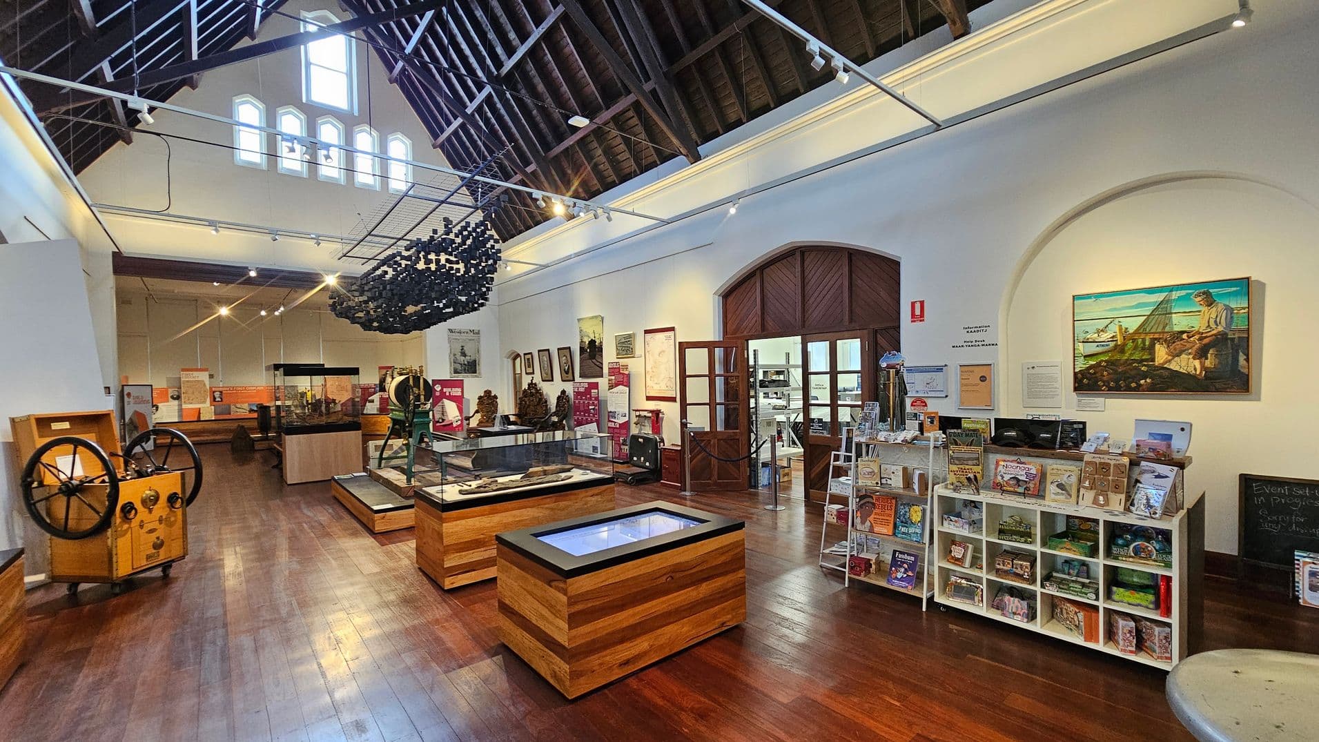A museum interior with a high, dark wooden ceiling and polished wooden floors. In the centre, there are display cases with artefacts. On the right, a wall features a colourful painting above shelves filled with books and souvenirs. A large wooden door with glass panels is in the background, leading to another room. The left side has various historical machinery and informational displays. Bright lighting illuminates the space, highlighting the exhibits and decor.