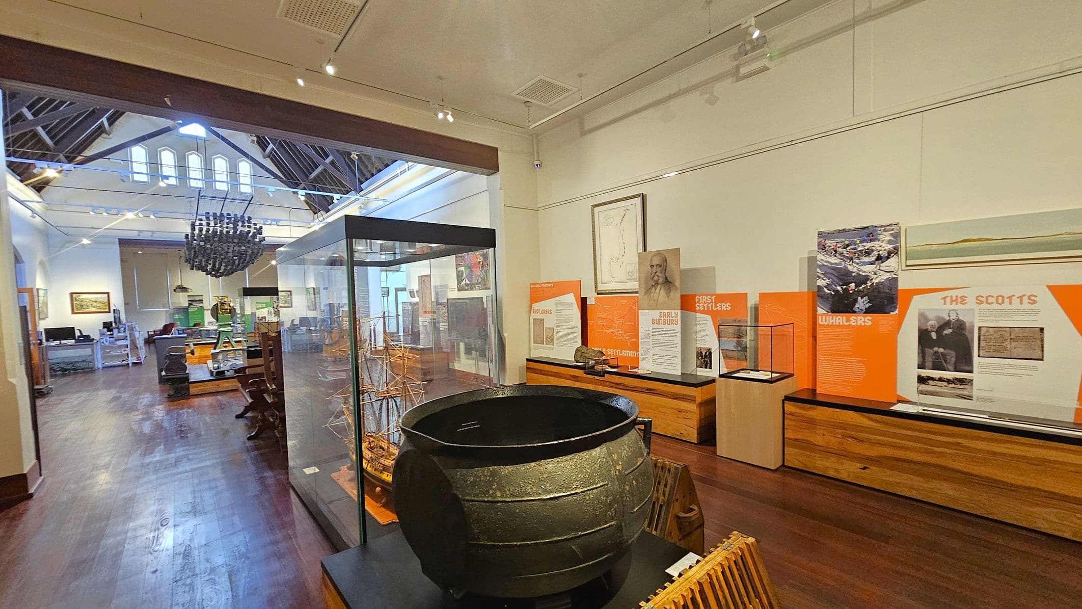 A museum interior with wooden flooring and a high, vaulted ceiling. On the right, there are informational displays with text and images about early settlers, whalers, and the Scotts. In the centre, a large black cauldron is displayed in front of a glass case containing a model ship. The background features more exhibits, including furniture and artefacts, with a large chandelier hanging from the ceiling. The room is well-lit with spotlights.