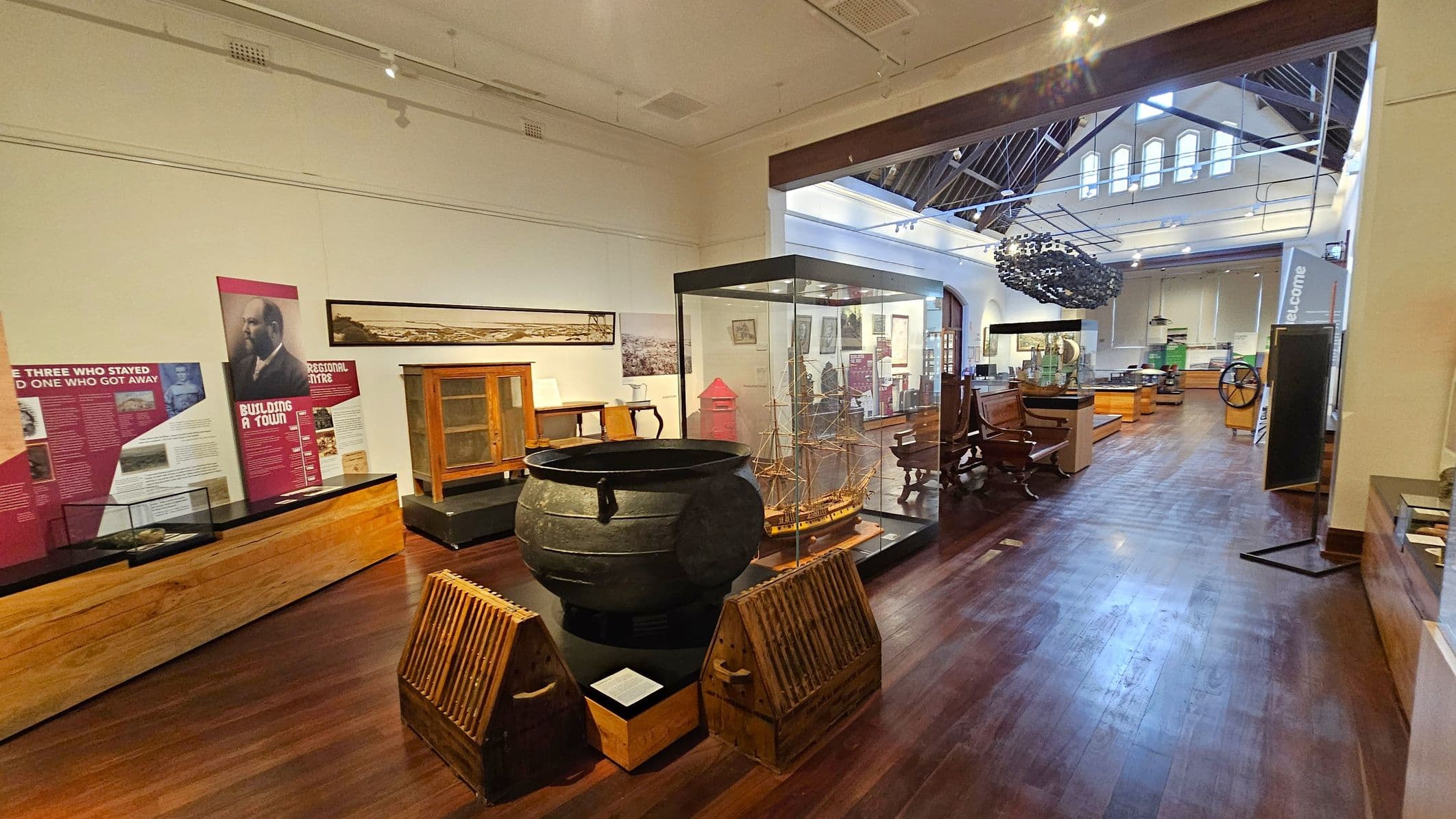 A museum interior with wooden flooring and a high ceiling featuring exposed beams. On the left, there are informational panels and a wooden display case. In the centre, a large black cauldron is flanked by wooden crates, and a model ship is encased in glass. Towards the back, various artefacts and furniture are displayed, including a large ship's wheel. The space is well-lit with natural light from high windows.