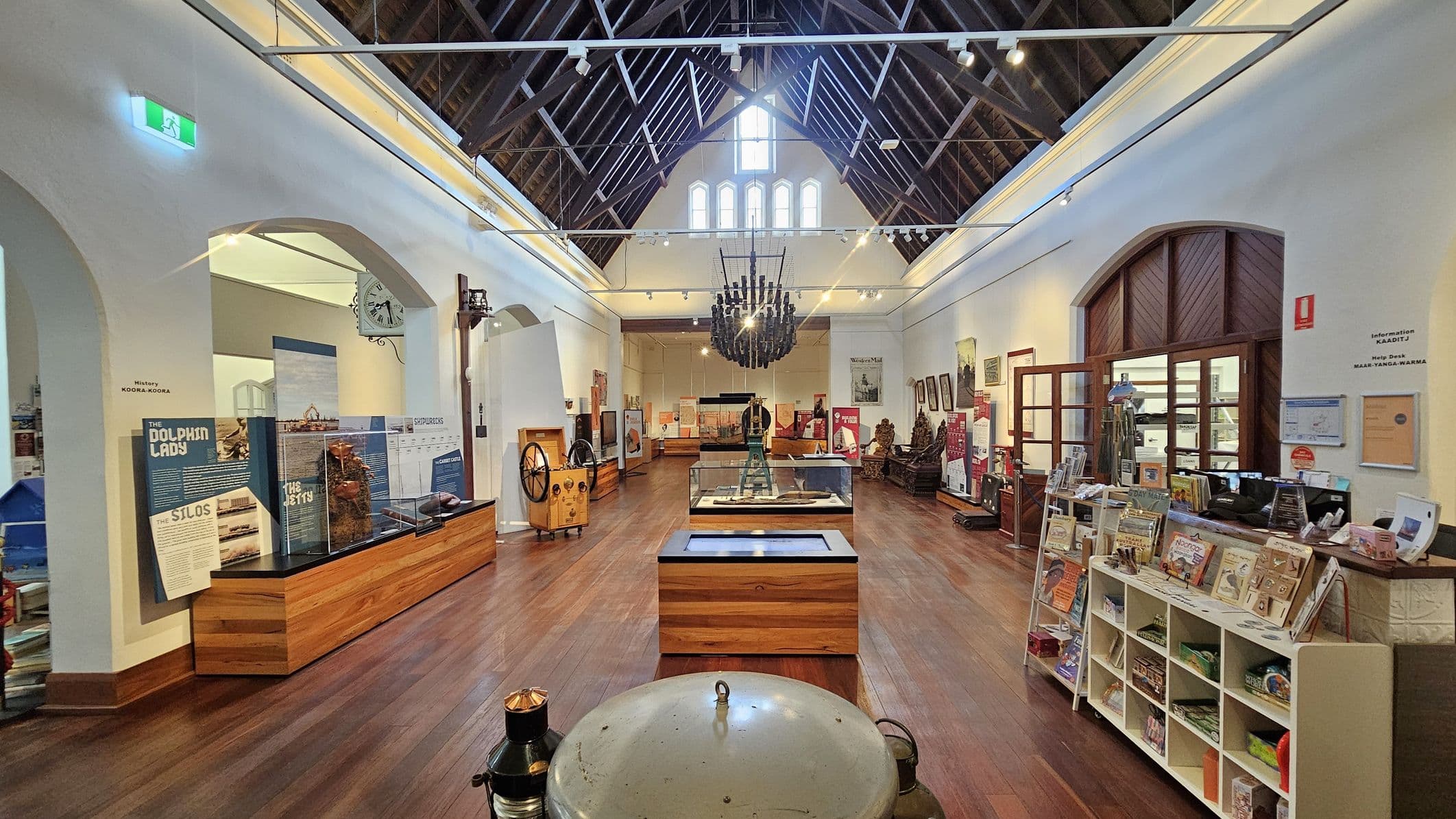 A museum interior with a high, wooden-beamed ceiling. In the centre, a glass display case sits on a wooden base, surrounded by various exhibits. On the left, informational panels and artefacts are displayed, including a clock mounted on the wall. On the right, a wooden shelf holds books and souvenirs. The floor is wooden, and a large chandelier hangs from the ceiling. Arched doorways and framed pictures are visible along the walls.