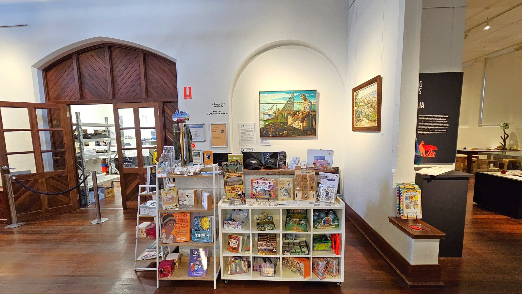 A room with wooden flooring and a high ceiling. On the left, there is a large wooden door with glass panels, leading to a storage area with shelves. In the centre, a display stand holds various books, souvenirs, and informational materials. Above the stand, a painting is mounted on the wall. On the right, another painting is visible, and a black panel with text and a red graphic is partially seen. A small shelf holds additional books.