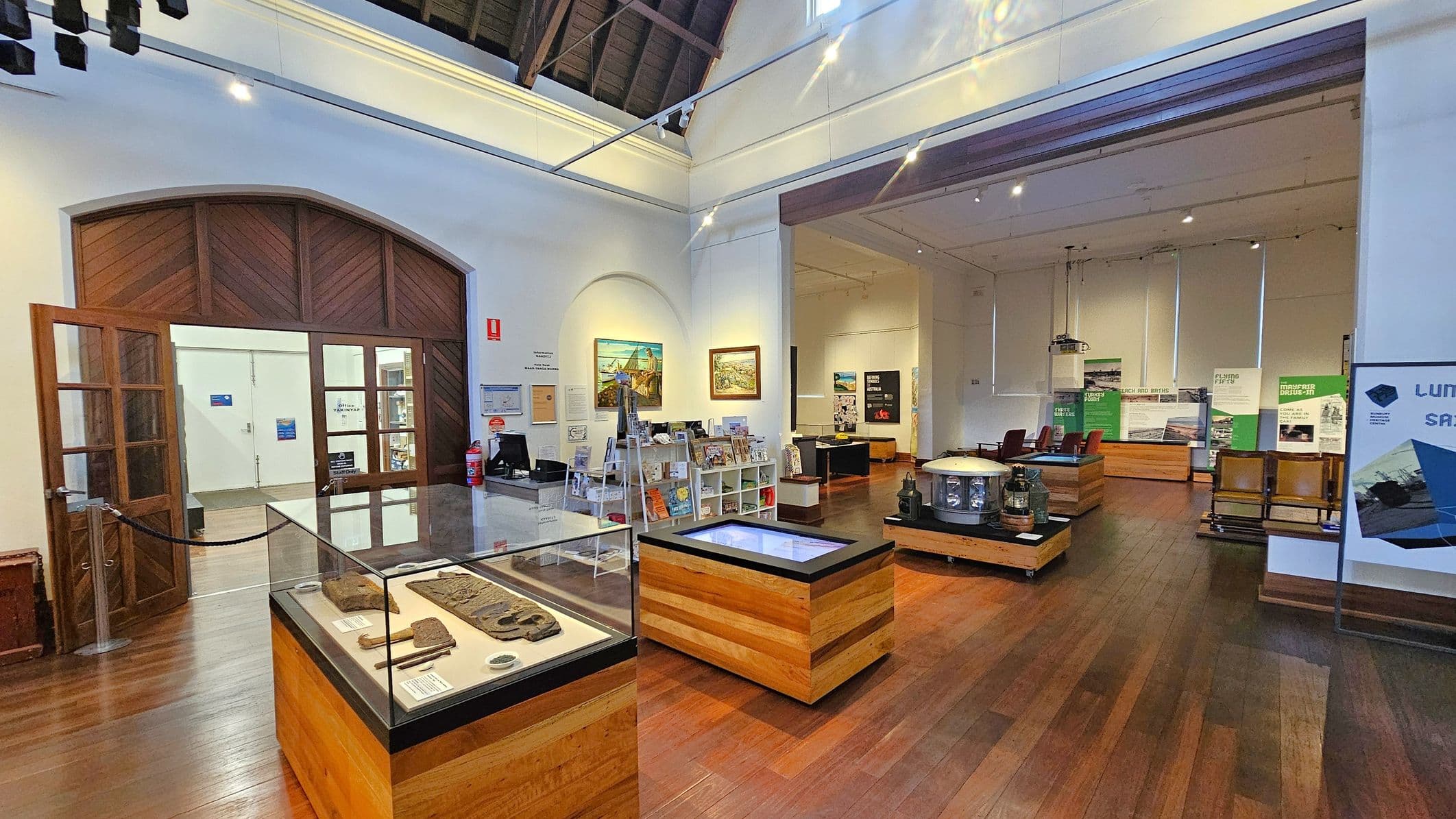 A museum interior with wooden flooring and a high ceiling. On the left, a large wooden door with glass panels leads to another room. In the centre, glass display cases showcase artefacts. The right side features informational panels and exhibits, with a few chairs and additional display cases. The walls are adorned with paintings and informational posters. A fire extinguisher is mounted on the wall near the door.