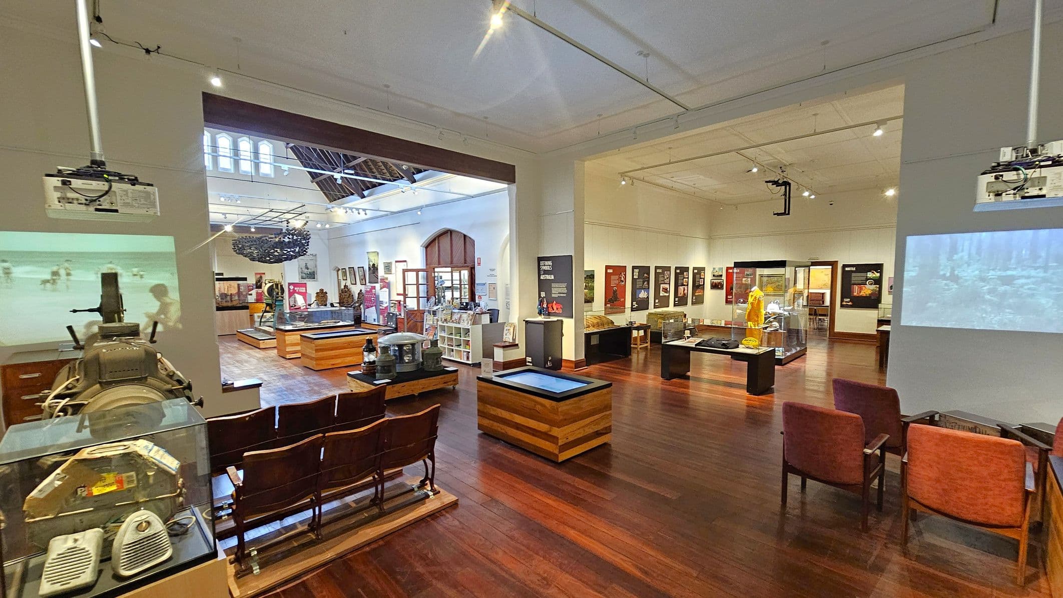 A museum interior with wooden flooring and a high ceiling. On the left, there are vintage cinema seats and a projector displaying an image on the wall. In the centre, various exhibits are displayed on wooden tables, with informational panels on the walls. The right side features more seating and a glass display case with a mannequin in a yellow outfit. The background shows a large arched window and additional exhibits, with bright lighting illuminating the space.
