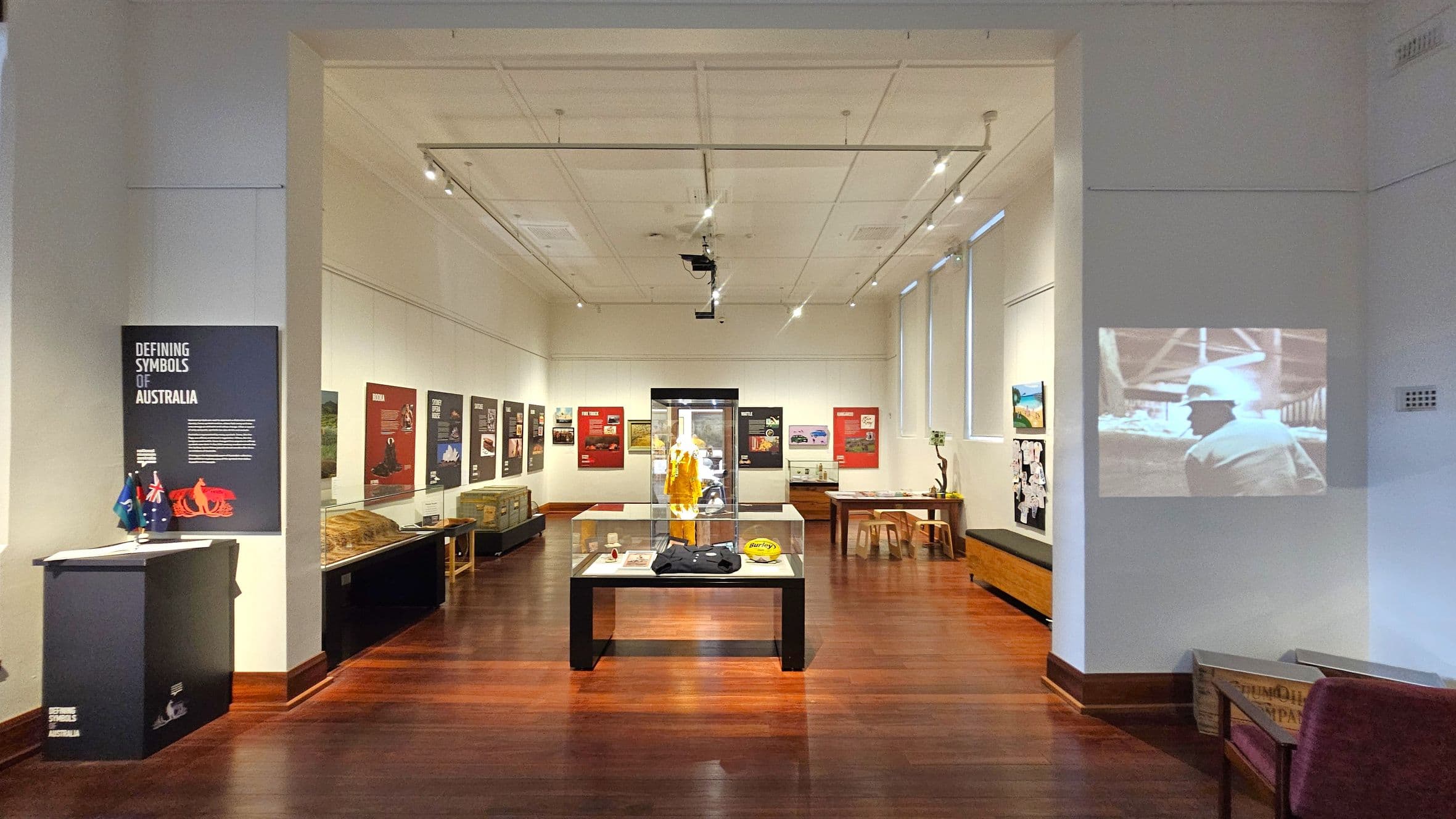 A museum exhibition room with wooden flooring and a white ceiling featuring track lighting. On the left, a display titled "Defining Symbols of Australia" with small flags and a red object. The centre has a glass case displaying a yellow garment and other items. The right wall has a projected black-and-white image. Various posters and artefacts line the walls, with benches and tables scattered throughout.