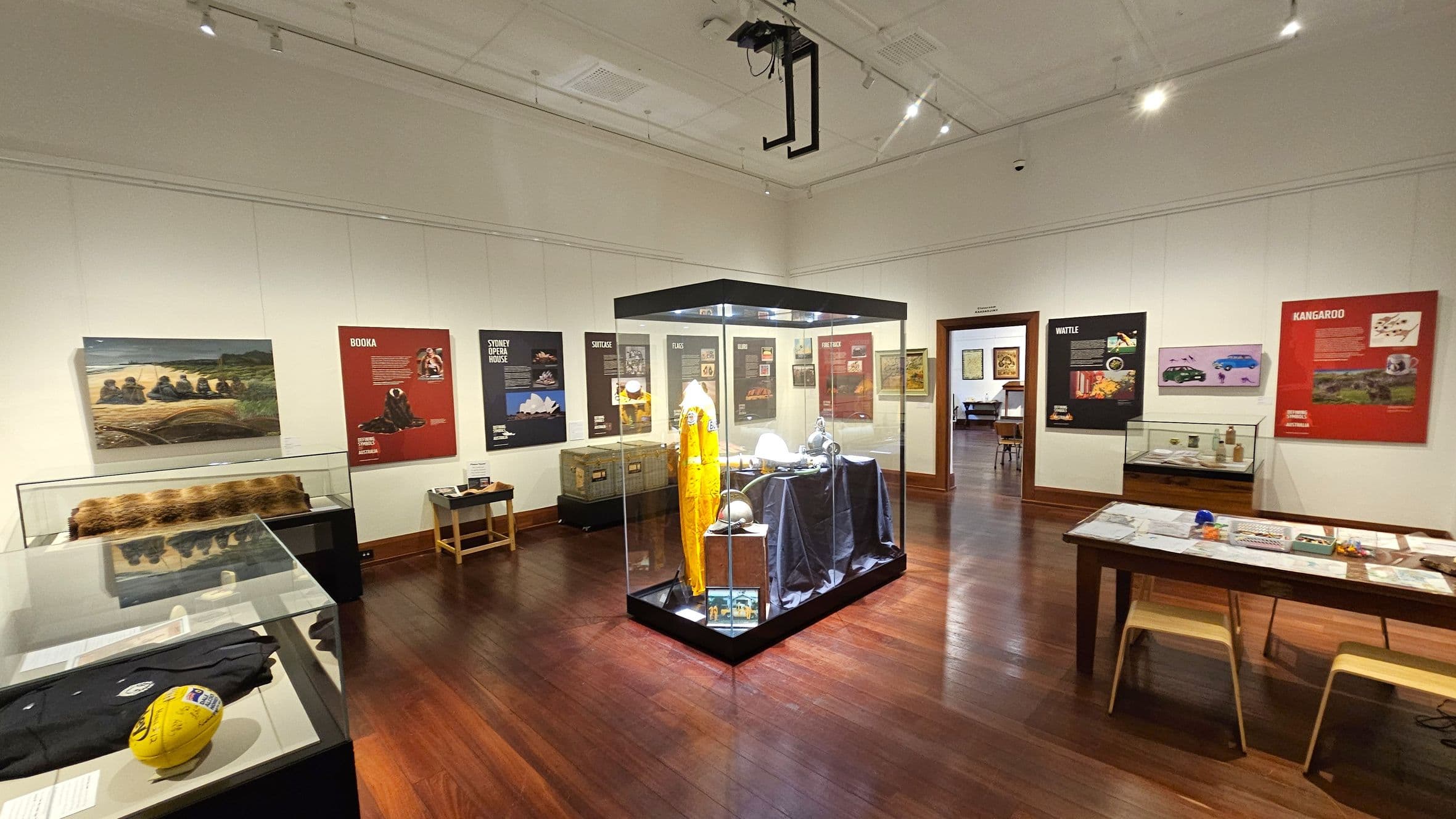 A museum exhibit room with wooden flooring and a white ceiling. In the centre, a glass display case contains a yellow suit and various objects. The walls are adorned with informational posters and images, including topics like "Booka," "Sydney Opera House," "Wattle," and "Kangaroo." Display cases on the left and right hold various artefacts. A doorway in the back wall leads to another room. A table with chairs is on the right, featuring papers and small items.