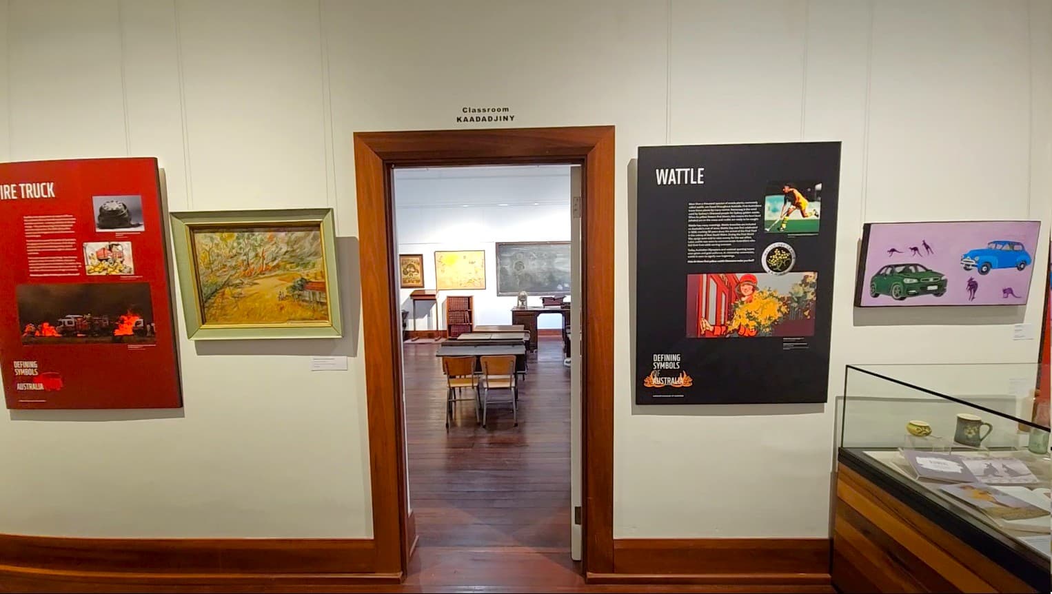 A museum room with a doorway in the centre leading to a classroom setup. On the left wall, there's a red panel with text and images about a fire truck, alongside a framed painting of a landscape. On the right wall, a black panel titled "Wattle" features text and images, with a colourful artwork of cars and kangaroos nearby. A glass display case is partially visible on the far right. The floor is wooden, and the ceiling is not visible.