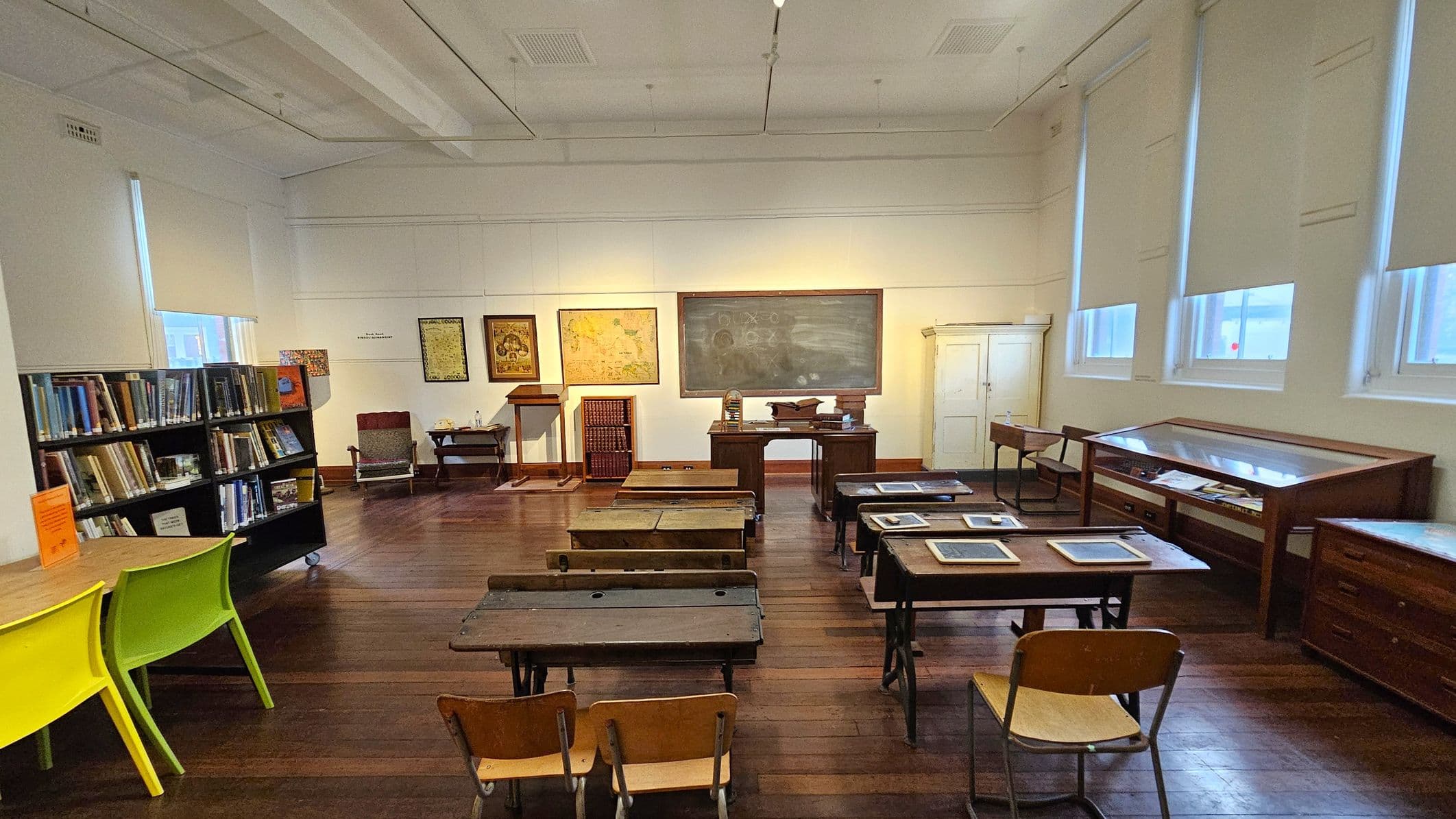 A vintage classroom setting with wooden desks and chairs arranged in rows on a wooden floor. At the front, there's a large chalkboard with drawings, flanked by framed maps and a lectern. To the left, bookshelves filled with books and two bright green chairs are visible. On the right, display cases with documents and a white cupboard are against the wall. The ceiling is high with visible beams, and windows with blinds allow natural light to enter.