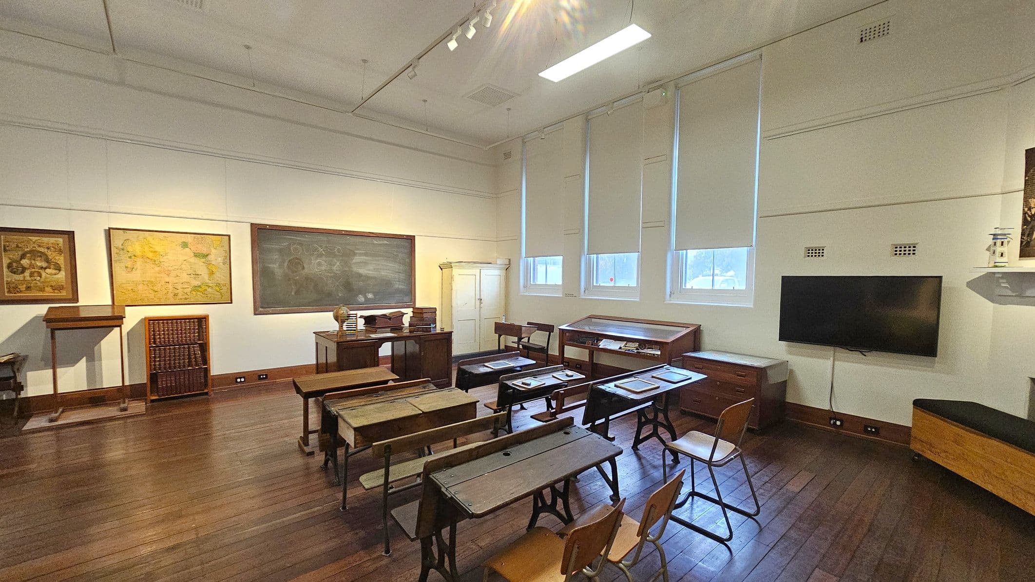 A vintage classroom with wooden desks and chairs arranged in rows on a polished wooden floor. On the left, there are framed maps and a bookshelf. In the centre, a large chalkboard with writing is mounted on the wall, accompanied by a teacher's desk with books. The right side features a modern flat-screen TV and display cabinets beneath windows with blinds. The ceiling is high with track lighting and a fluorescent light fixture.