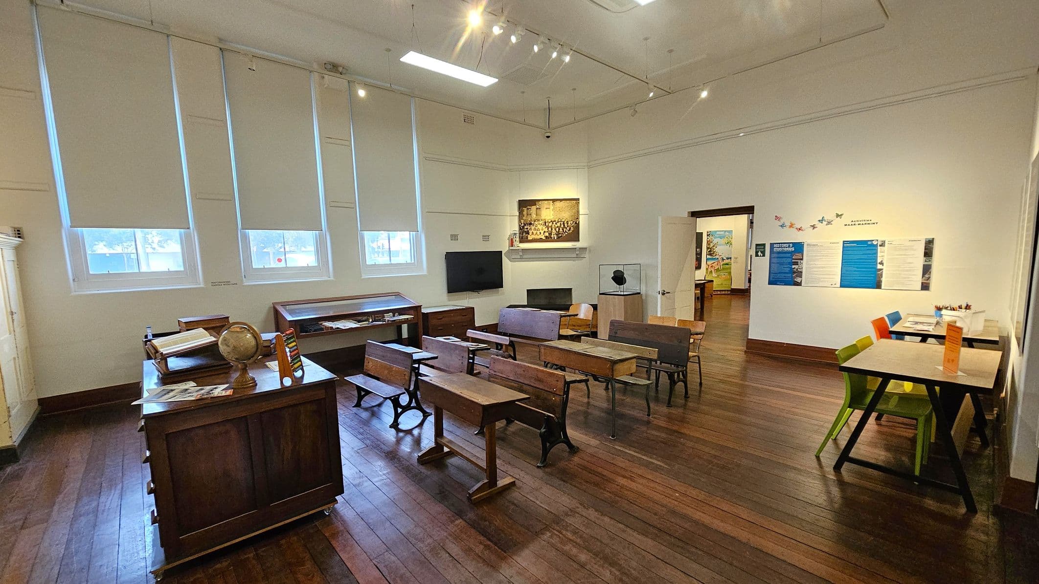 A room with wooden flooring and a high ceiling. On the left, there are vintage wooden school desks and a teacher's desk with a globe and books. Three windows with blinds are above the desks. In the middle, more wooden desks are arranged in rows. On the right, a modern table with colourful chairs is against the wall, which has educational posters and a butterfly decoration. An open door leads to another room in the background.