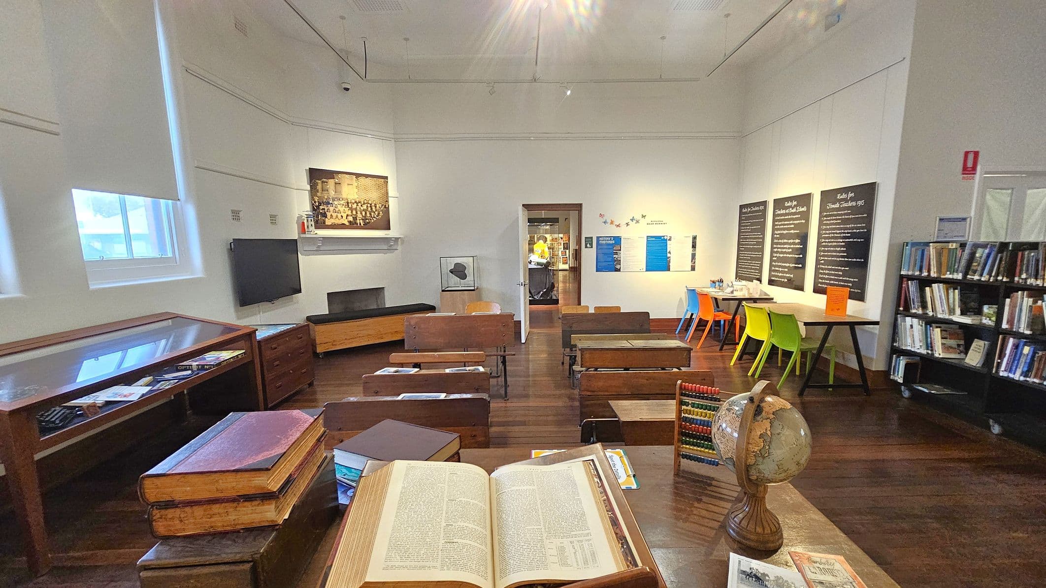 A room with wooden flooring and a high ceiling. In the foreground, an open book, a globe, and an abacus are on a desk. To the left, a display case and a wall-mounted TV are visible. In the centre, rows of vintage wooden desks face a doorway leading to another room. On the right, colourful chairs are around a table beneath informational boards. A bookshelf filled with books is against the right wall. The room is well-lit with natural light from windows on the left.