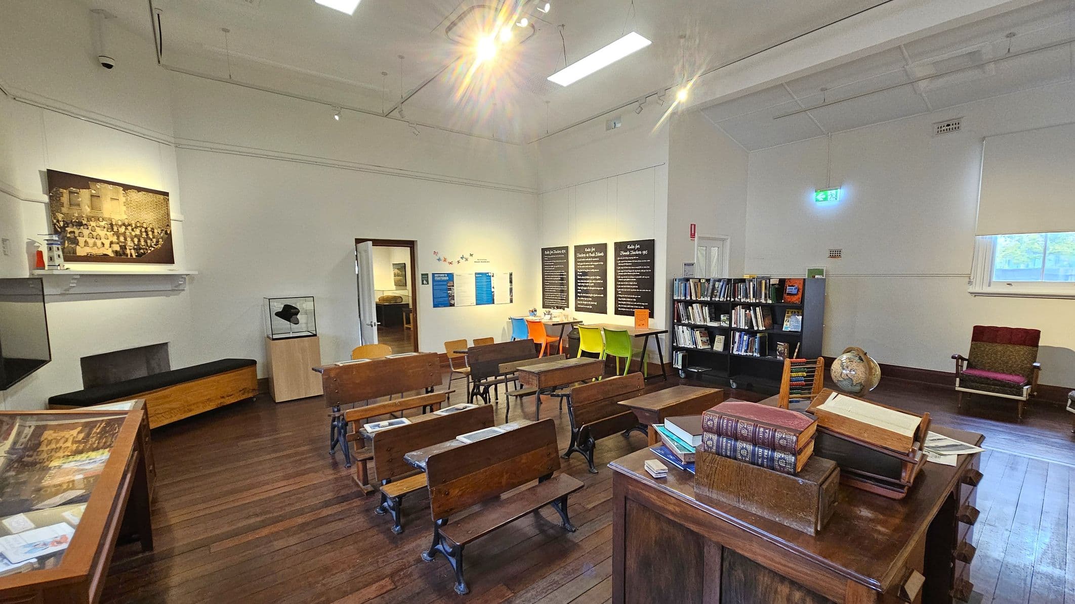 A room with wooden flooring and a high ceiling. On the left, there's a large black-and-white photograph above a bench and a display case. In the centre, rows of vintage wooden desks face a wall with educational posters and colourful chairs. On the right, a bookshelf is filled with books, and a globe sits on a table with stacked books. A single armchair is near a window with a blind. The room has a mix of historical and educational elements.