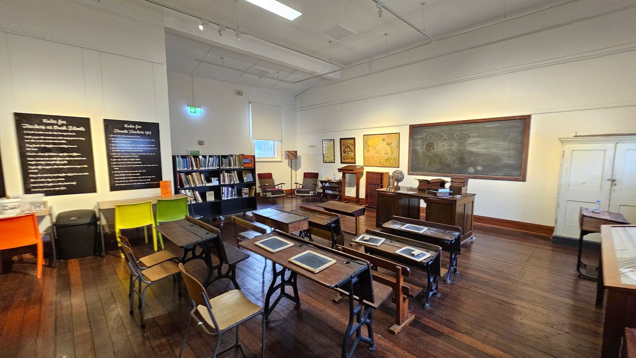 A vintage classroom with wooden desks and chairs arranged in rows. On the right, a large chalkboard with writing is mounted on the wall. In the background, a teacher's desk with a globe and books is positioned near a lectern. The left side features colourful chairs and informational boards on the wall. Bookshelves filled with books are visible near the back wall. The floor is wooden, and the ceiling has recessed lighting.