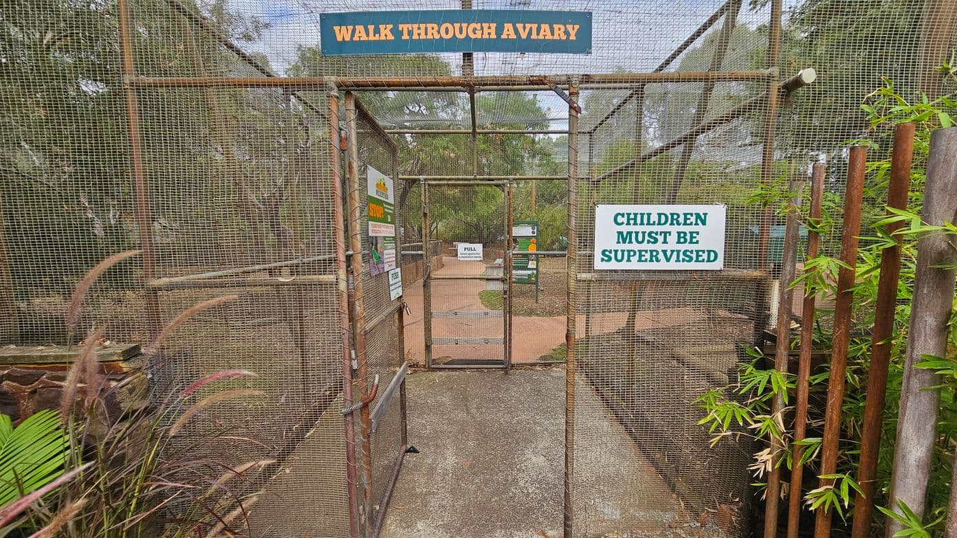 A metal mesh gate labelled "WALK THROUGH AVIARY" at the top. Signs on the gate read "CHILDREN MUST BE SUPERVISED" and "PULL". The path beyond the gate is paved and surrounded by greenery, including trees and plants. The ground is concrete, and there are wooden poles with green foliage on the right side. The background shows more signs and a continuation of the path.
