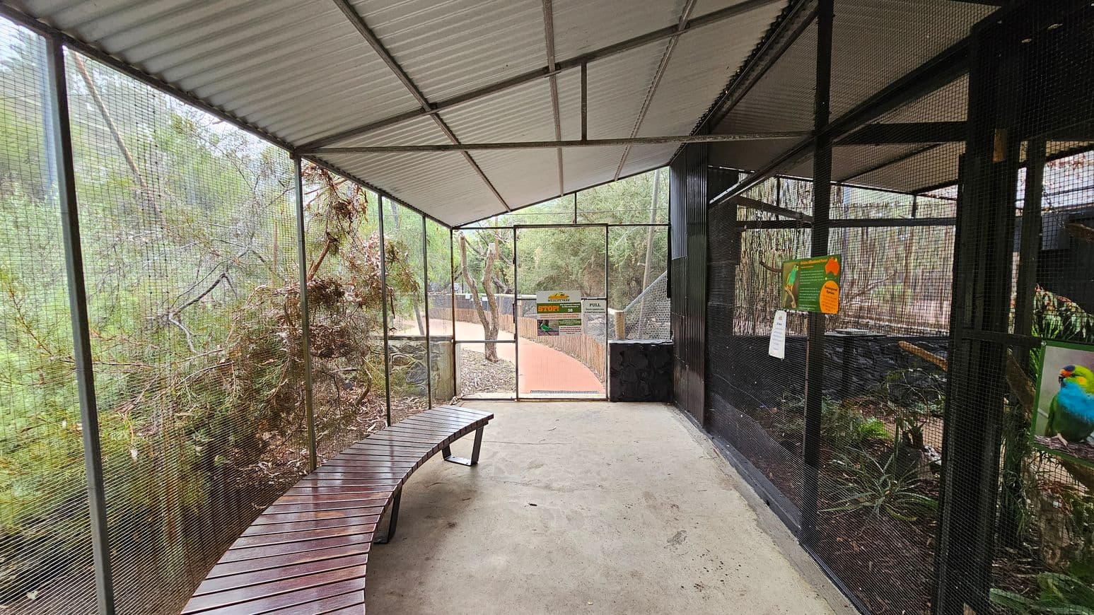 A covered walkway with a metal mesh enclosure on both sides. On the left, there's a curved wooden bench and dense foliage visible through the mesh. On the right, signs and a colourful bird image are attached to the mesh. The floor is concrete, and the ceiling is corrugated metal. In the centre, a gate with signs leads to a path outside, surrounded by trees.