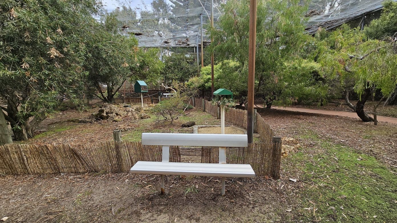 A white bench is positioned in the foreground, facing a fenced garden area. The garden is filled with trees and shrubs, with a few small green-roofed structures visible. The ground is covered with mulch and patches of grass. In the background, a netted canopy is stretched overhead, supported by tall poles. A red pathway runs along the right side, partially obscured by trees.