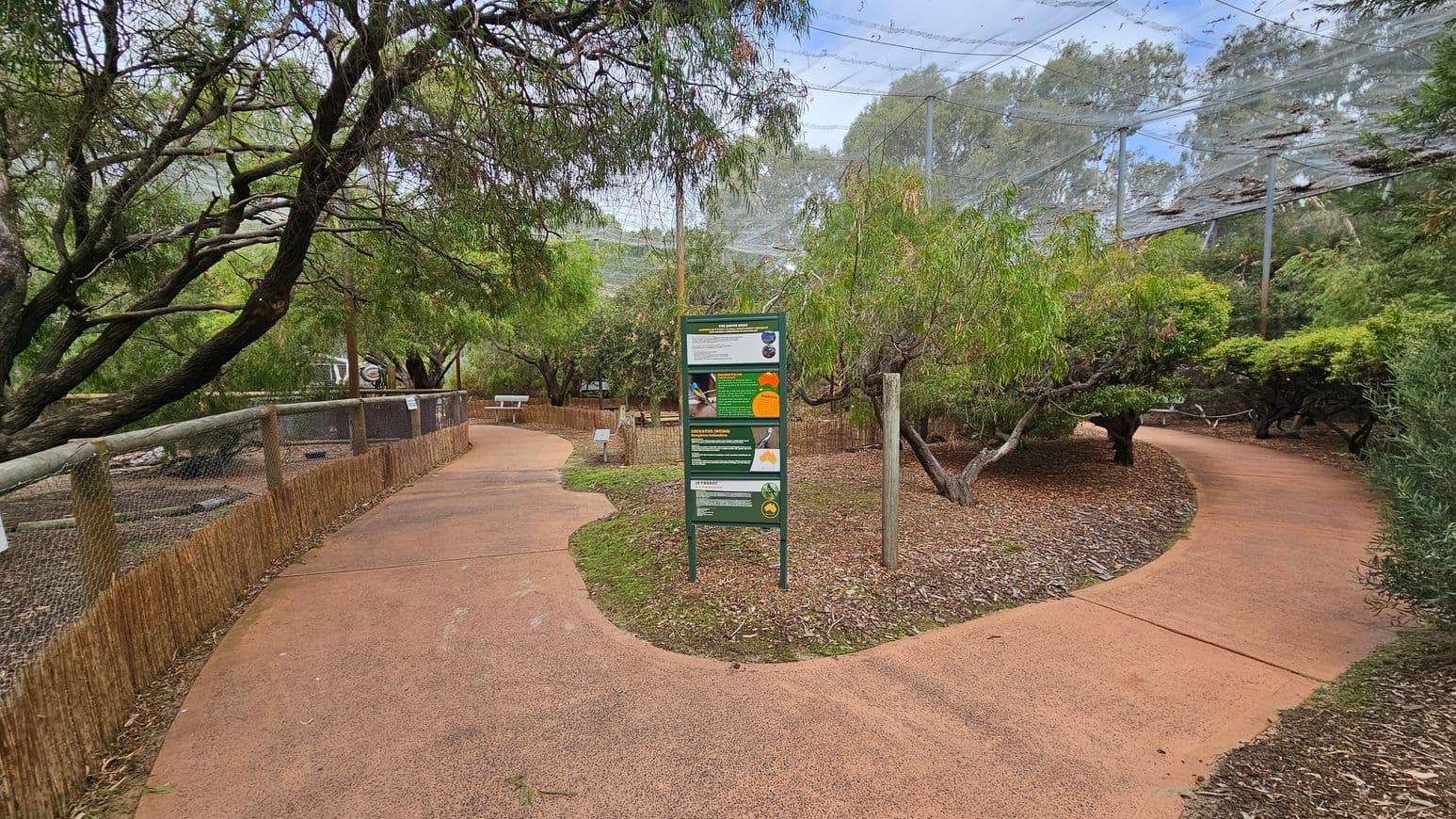 A pathway in a park or zoo with two diverging paths. On the left, a fenced area with netting overhead and trees lining the path. In the centre, a green informational sign with text and images. On the right, another path curves through more trees and foliage. The ground is covered with a reddish-brown surface, and there are wood chips and grass around the paths. The area is enclosed with netting above, and there are benches and tables visible in the background.